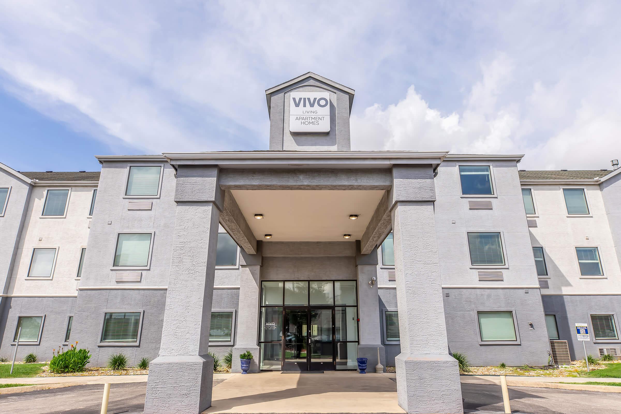 Exterior view of the VIVO Living Apartment Homes building, featuring a modern design with a prominent entrance under a triangular roof. The facade is light gray with large windows, and landscaping is visible at the front. The sky is partly cloudy, contributing to a bright atmosphere.