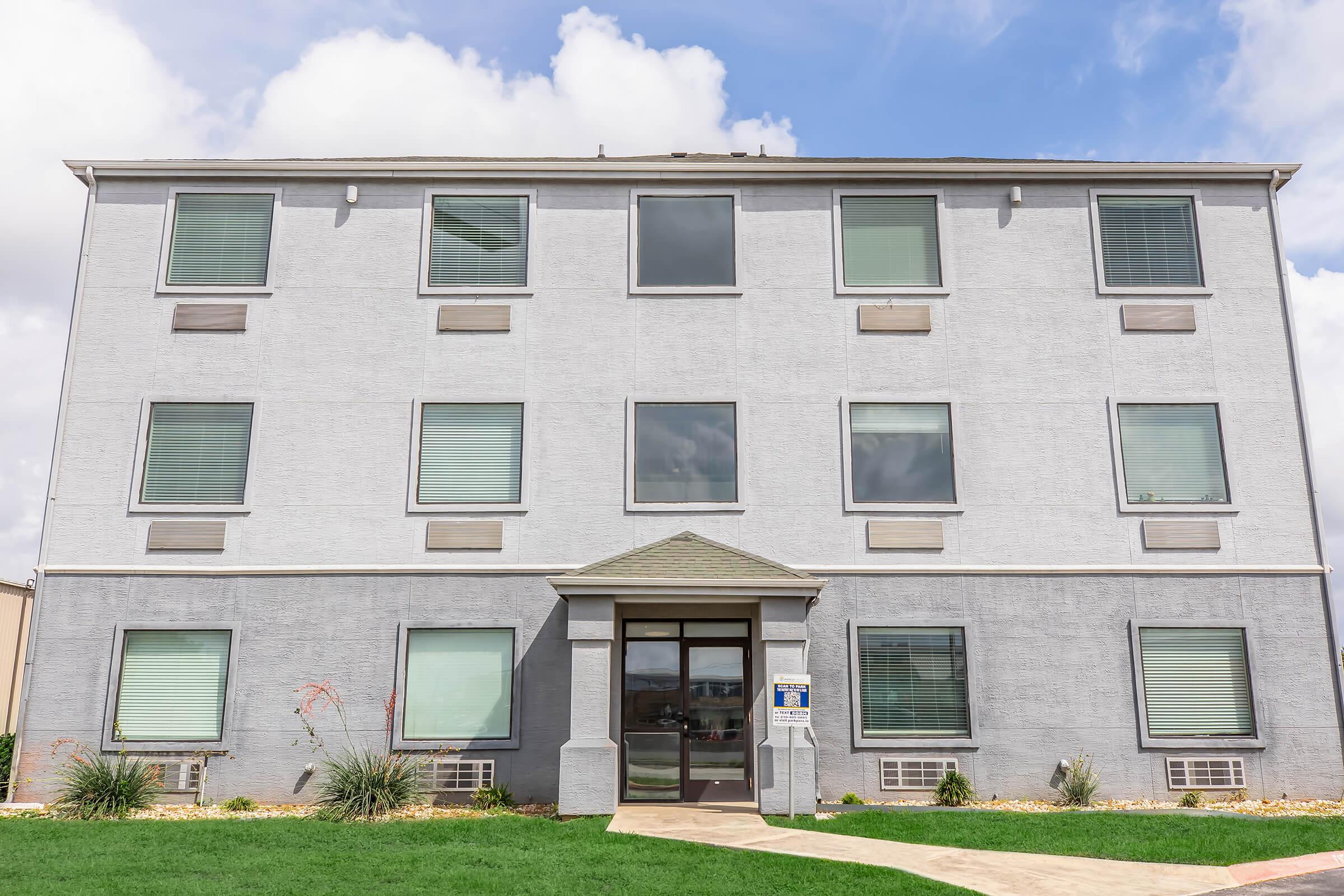 A three-story, gray exterior building with multiple windows. The front entrance features a covered porch and is flanked by windows. Lush green grass and decorative landscaping surround the building under a partly cloudy sky.