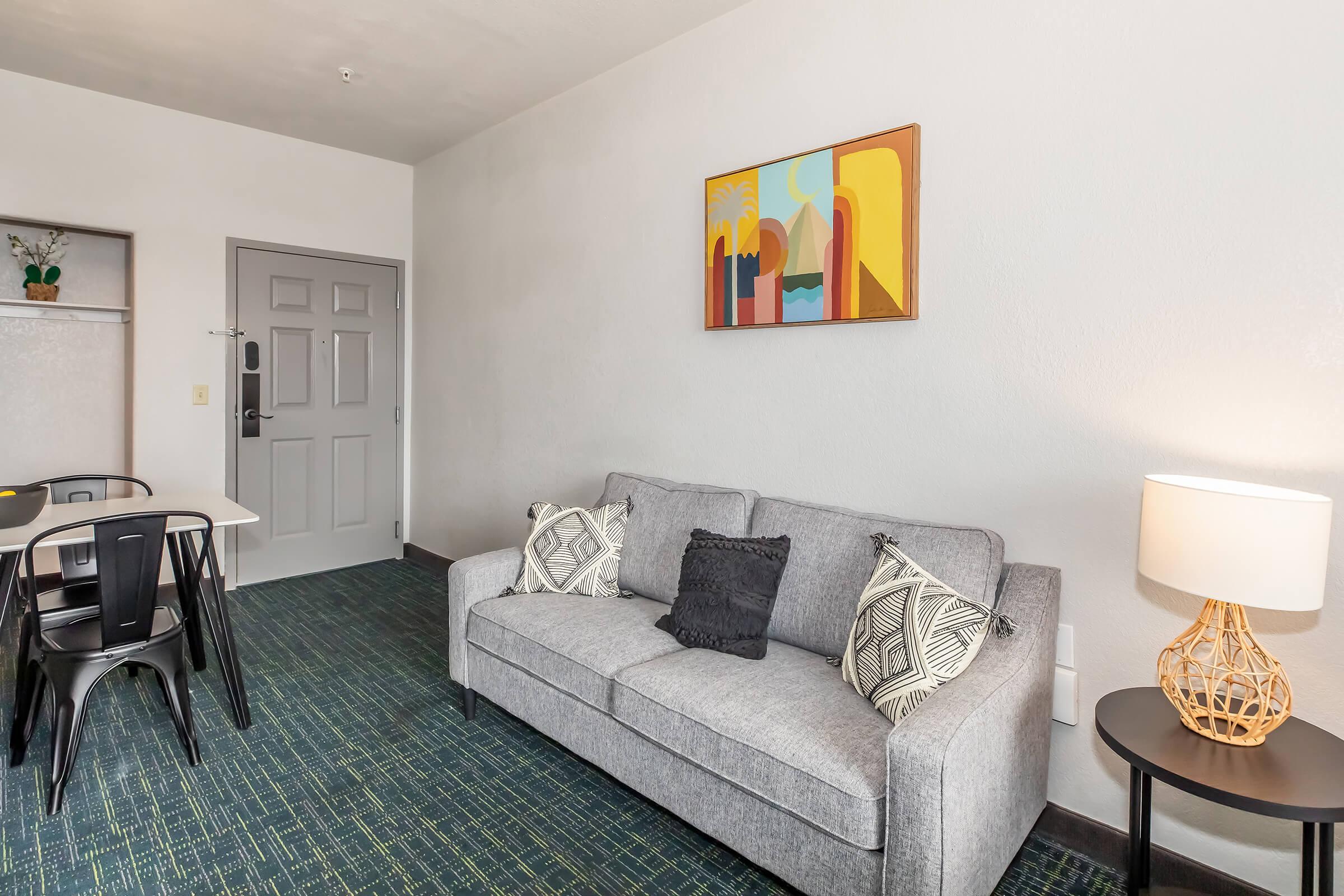 A cozy living area featuring a gray sofa with decorative pillows, a small dining table with black chairs, a modern lamp, and a colorful abstract painting on the wall. The floor is carpeted with green tones, and there is a door leading outside visible in the background.