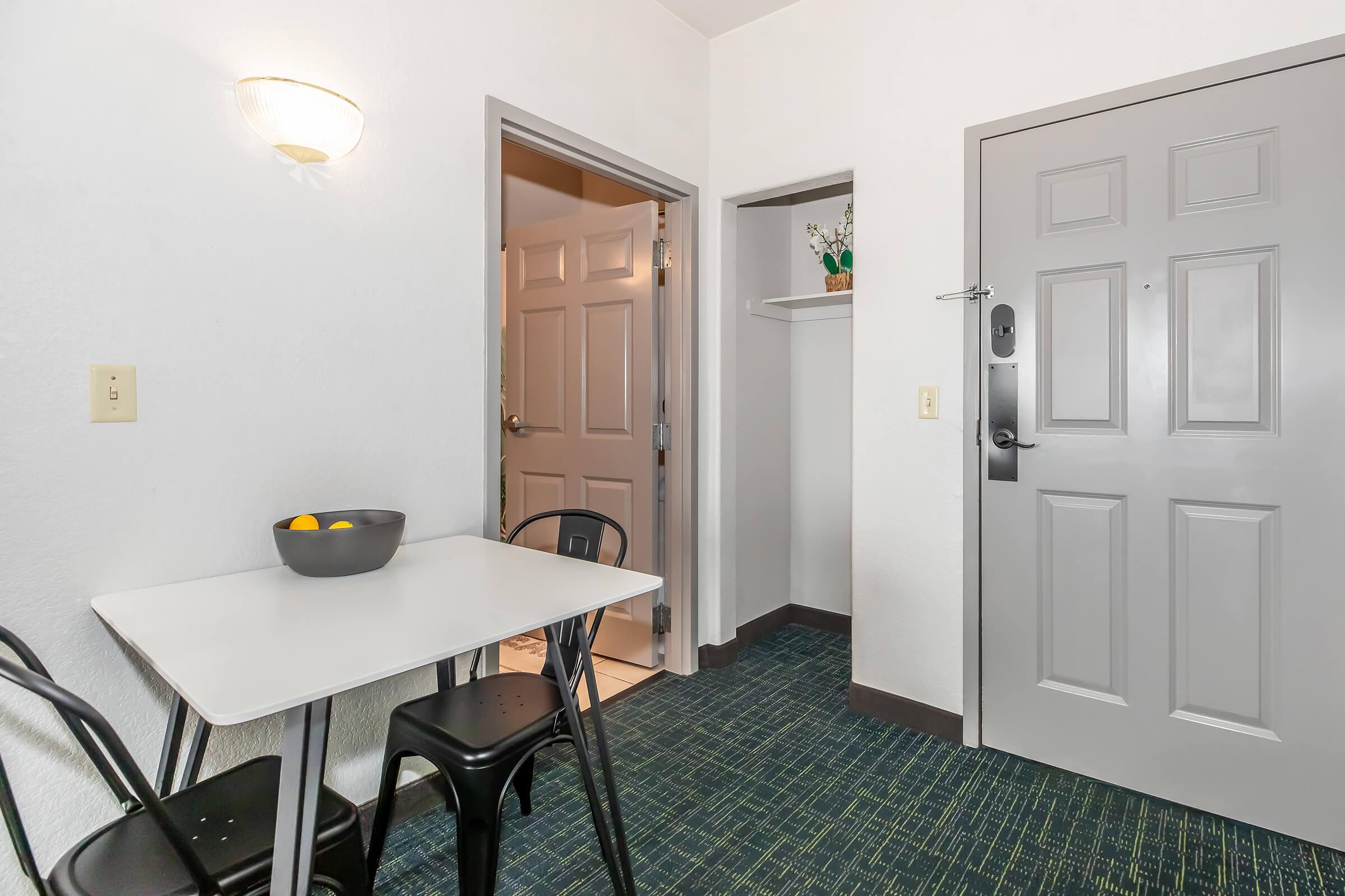 A small entryway featuring a table with a bowl of oranges, black chairs, and a closet door. The wall has a light fixture, and an entrance door is visible in the background. The flooring has a green pattern, contributing to a simple and tidy living space atmosphere.