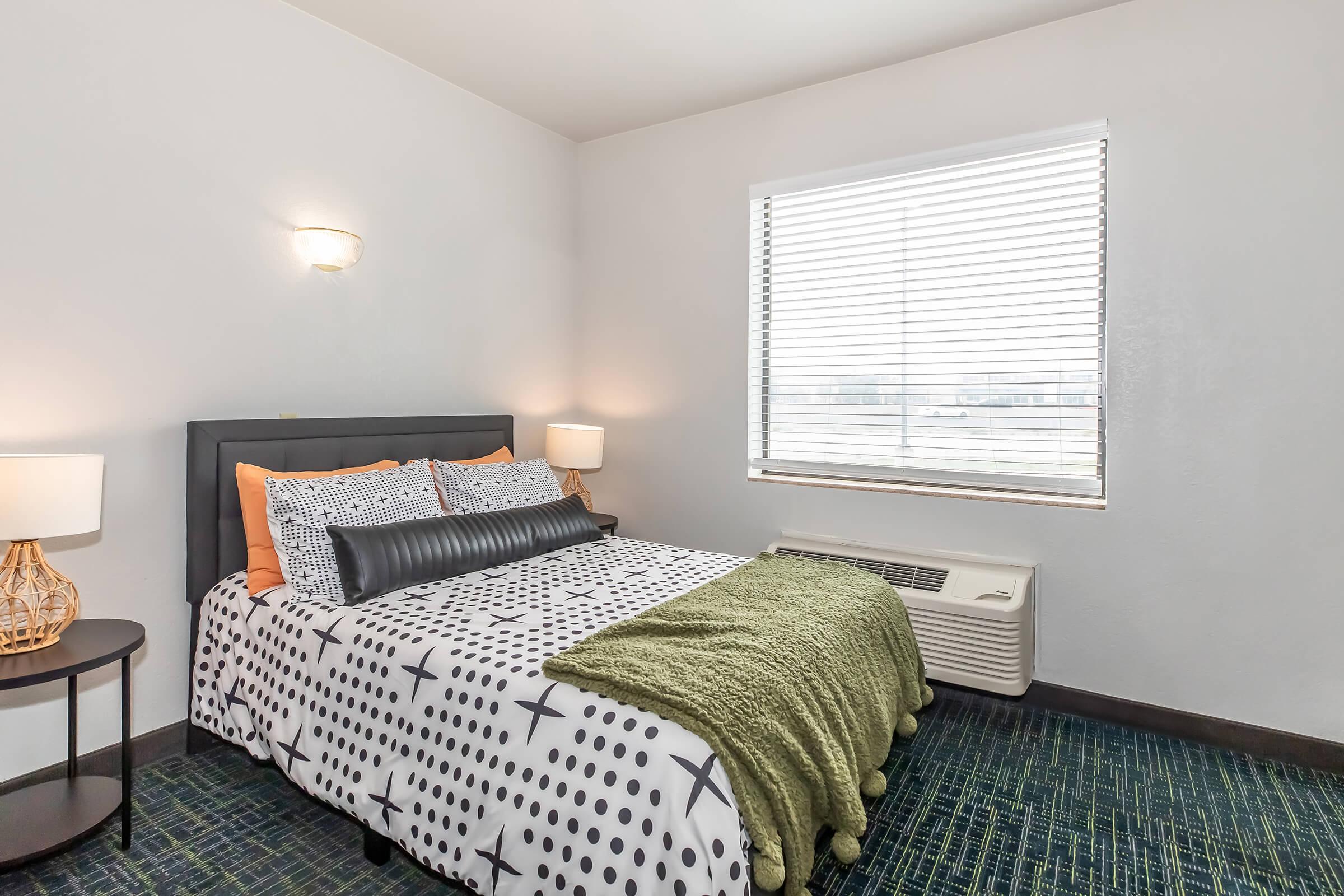 Cozy bedroom featuring a bed with patterned bedding and a dark headboard. There are two lamps on either side of the bed, and a stylish nightstand. A green throw blanket is draped over the foot of the bed. Natural light enters through a window with blinds, which overlooks a muted interior.