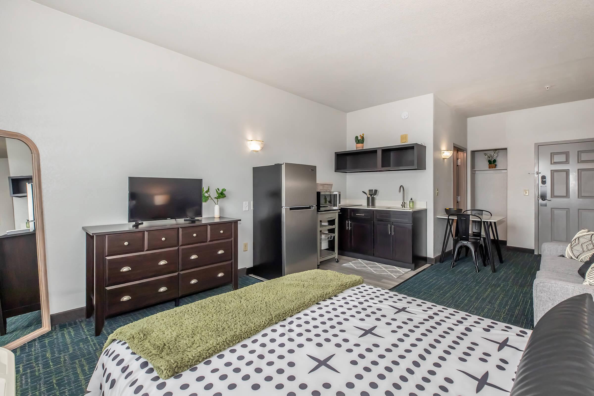 A spacious hotel room featuring a bed with a patterned blanket, a dresser, a television, a kitchenette with a refrigerator and microwave, a small dining table with chairs, and a cozy seating area. The room has light-colored walls and a carpeted floor, creating a welcoming atmosphere.