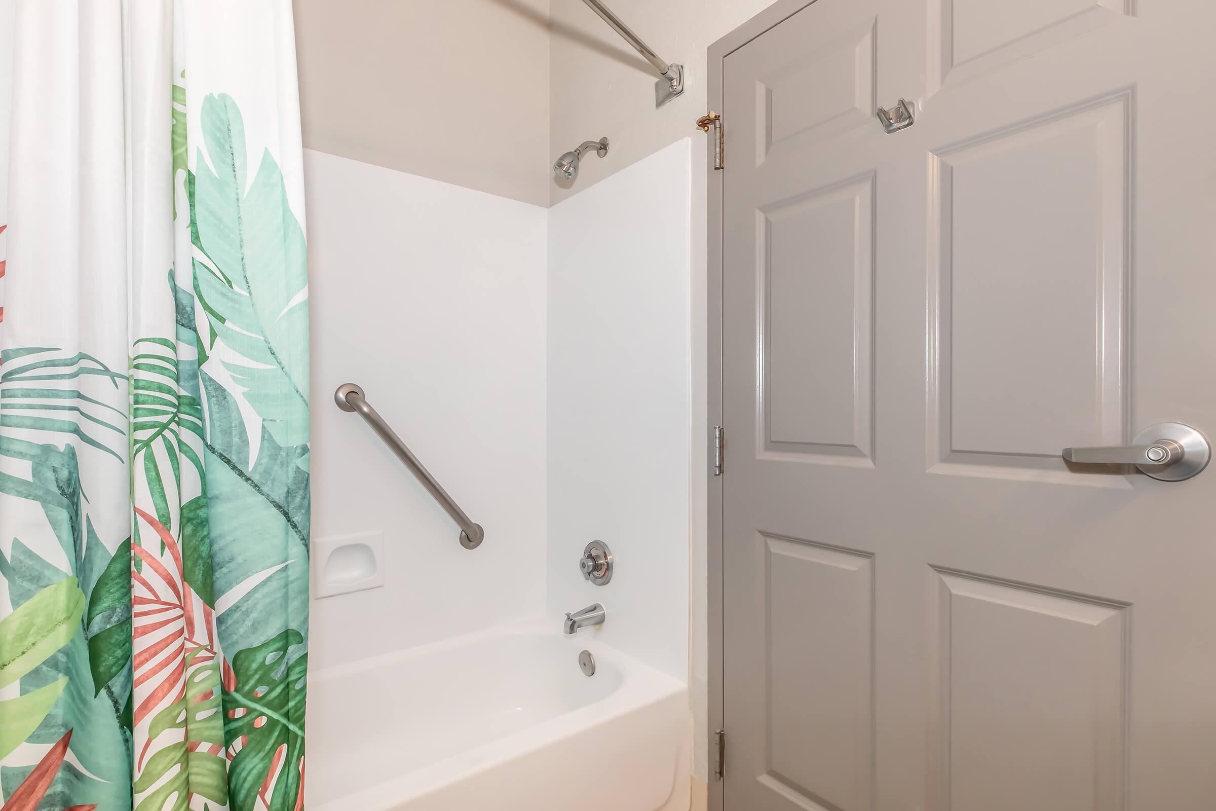 A clean bathroom featuring a shower-tub combination with a colorful tropical leaf shower curtain. There's a grab bar for safety and a gray door. The walls are plain white, and the overall ambiance is bright and inviting.