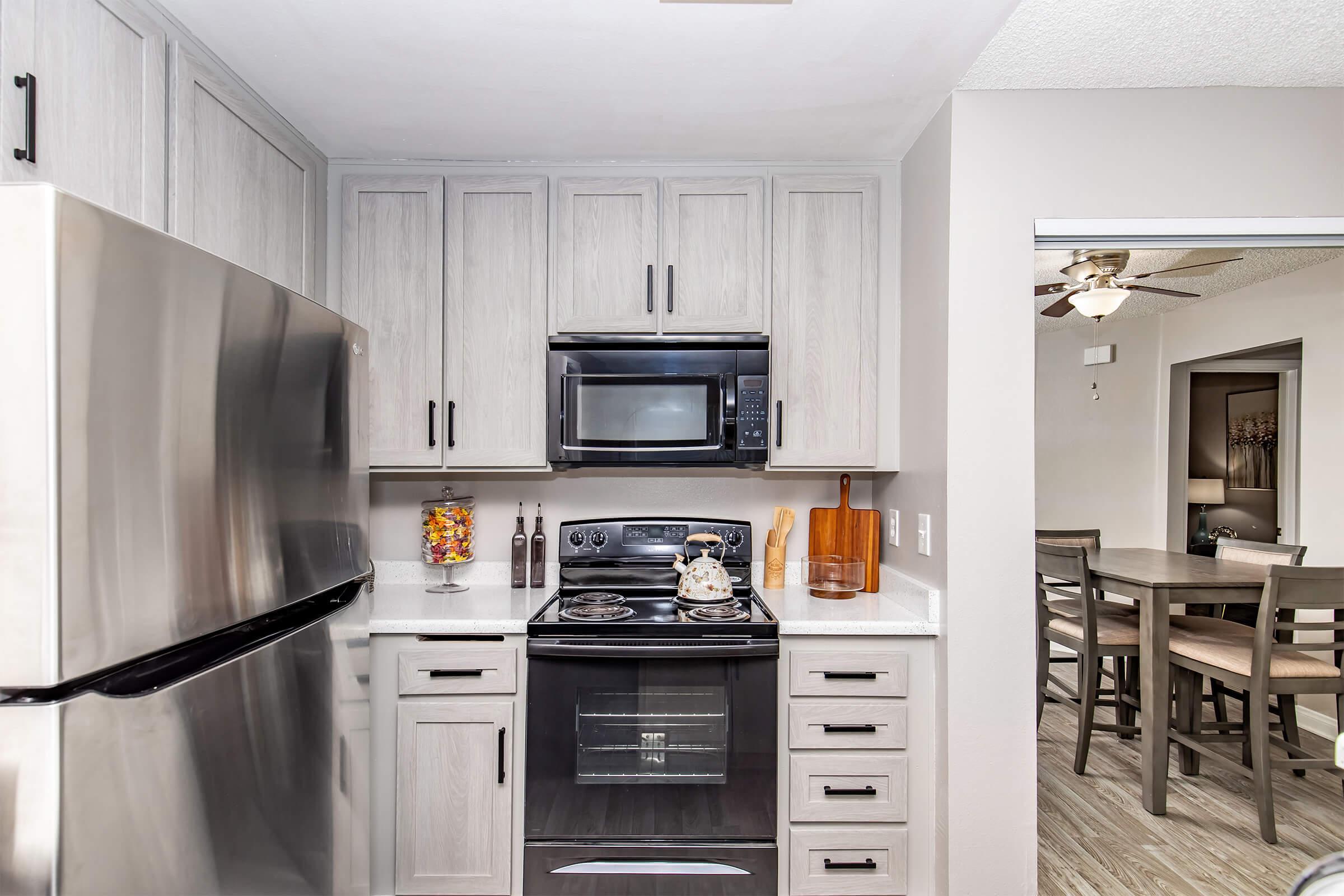 A modern kitchen featuring stainless steel appliances, including a refrigerator and microwave. The cabinetry is light-colored with a sleek finish, and the countertop is white. A small decorative element, such as a jar of colorful treats, is on the counter. In the background, a dining area is visible with a table and chairs.