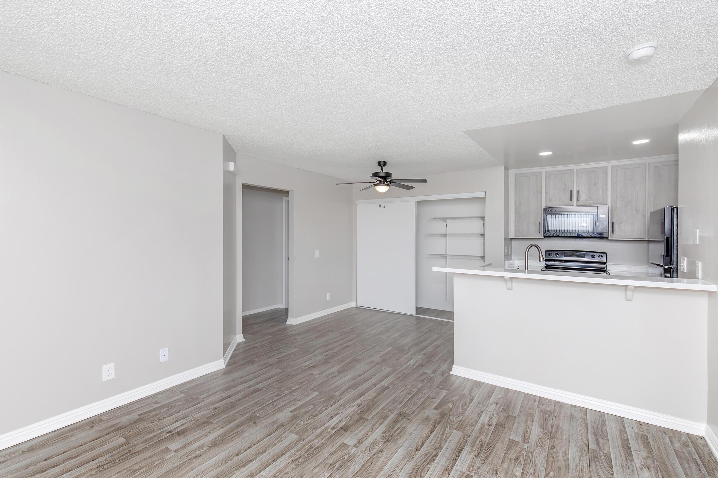 Modern, open-concept living space featuring light walls and wooden laminate flooring. A ceiling fan is mounted in the living area, which connects to a kitchen with a breakfast bar, cabinets, and appliances. The space is bright and uncluttered, with a doorway leading to another room.
