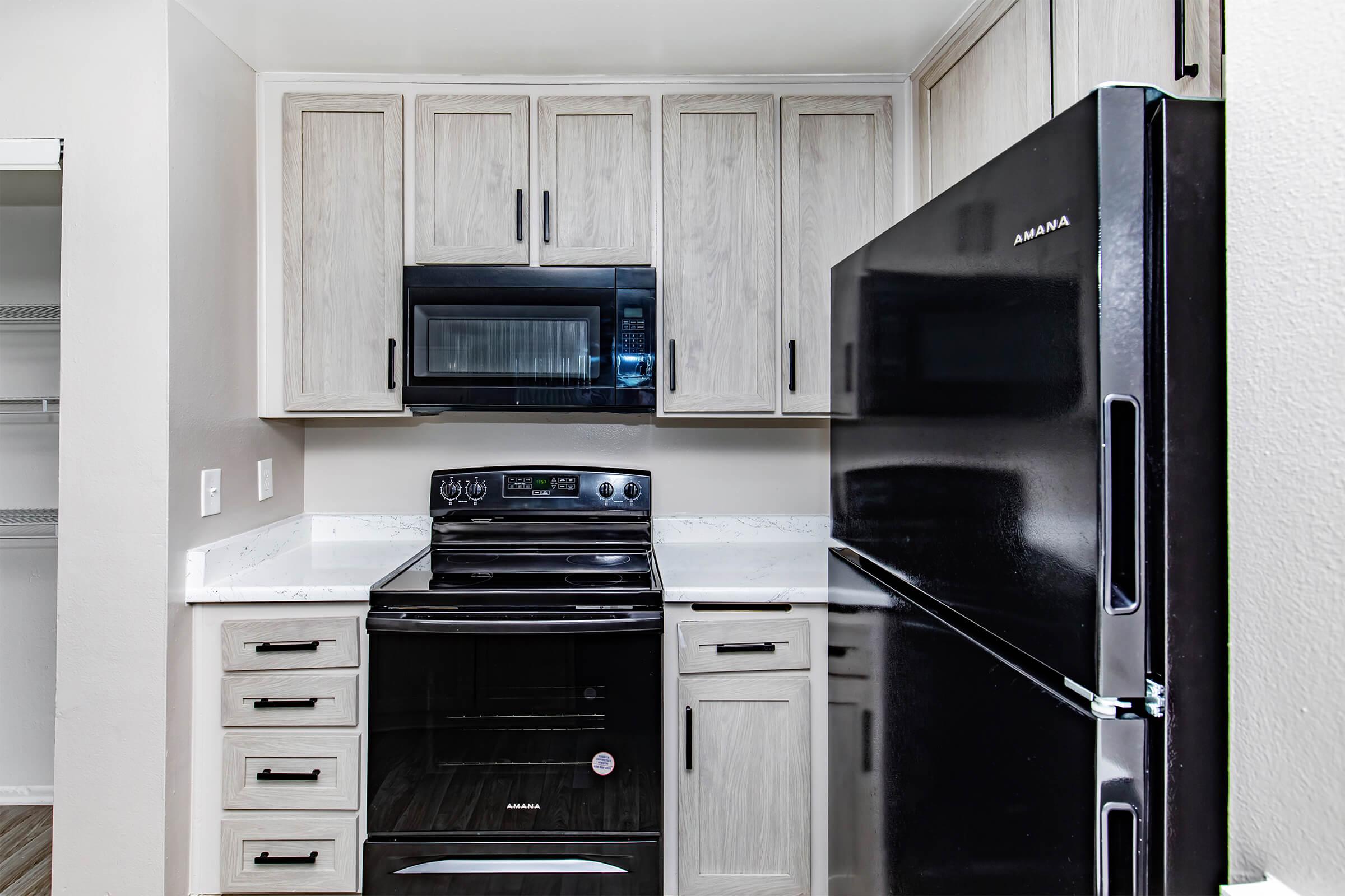 A modern kitchen featuring light wooden cabinets, a black stove, and a black refrigerator. The countertops are white, and there is a microwave above the stove. The walls are painted in a neutral color, enhancing the clean and contemporary look of the space.