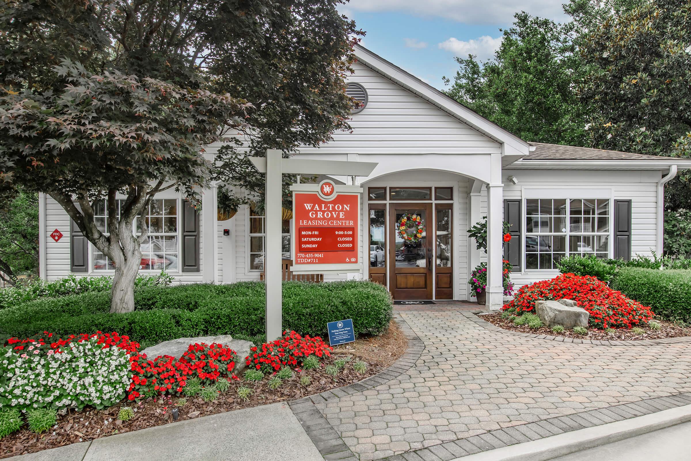 A charming white building with a peaked roof, surrounded by vibrant flower beds filled with red and white blooms. A sign for "Walton Grove Learning Center" is prominently displayed in front, detailing services offered. Lush green landscaping and trees enhance the welcoming atmosphere of the center.