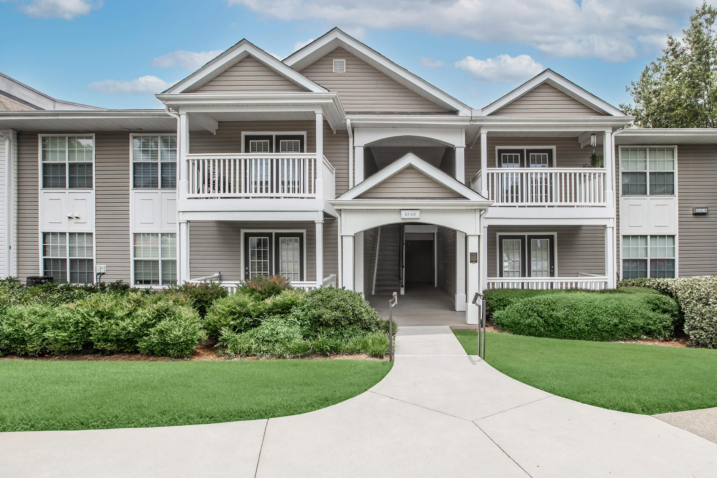 A well-maintained multi-unit residential building featuring a light gray exterior, white balconies, and landscaped green lawns. A pathway leads to the entrance, which has stairs on one side and a covered entryway. The sky is partly cloudy, adding to the serene atmosphere.