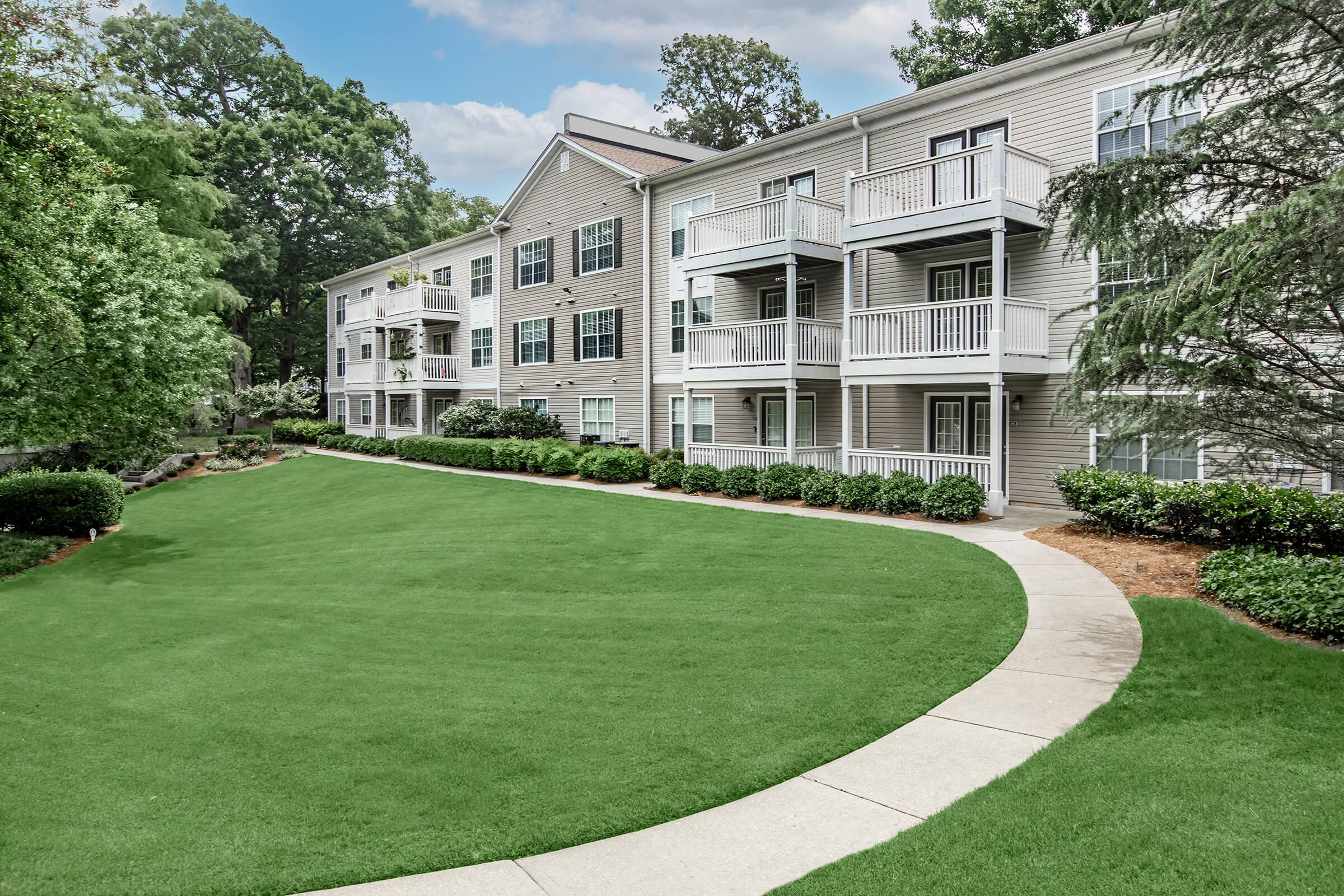 View of a residential complex featuring multiple apartment buildings with balconies, surrounded by well-maintained green lawns and landscaped walkways. The scene is set against a backdrop of trees and a partly cloudy sky, conveying a peaceful and inviting atmosphere.