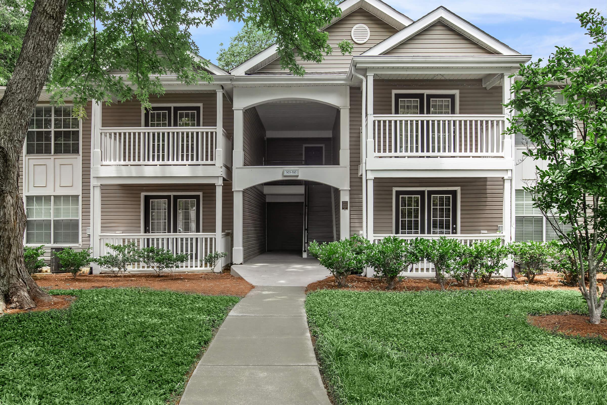 Two-story apartment building with a porch on each level, surrounded by green grass and shrubs. A paved walkway leads to the entrance, flanked by trees on either side. The building features a light brown exterior with white railings and trim, under a clear blue sky.