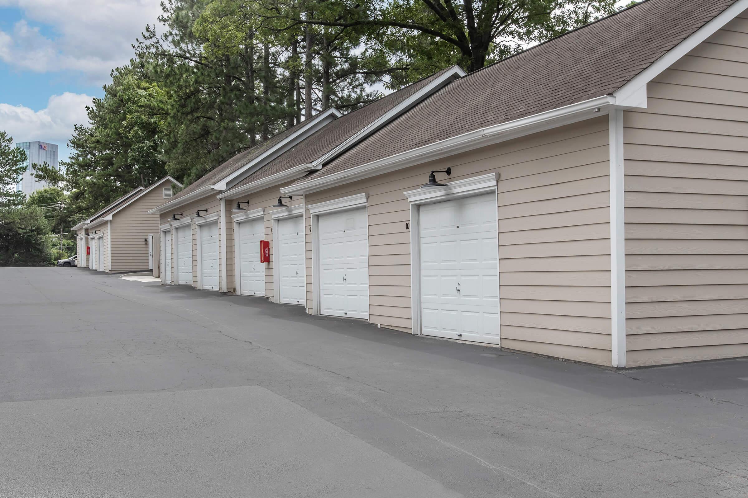 Row of beige garage doors lining a paved driveway, surrounded by trees. The garages have white doors and some are equipped with exterior lighting. The scene is set under a cloudy sky, adding a tranquil atmosphere to the neighborhood setting.