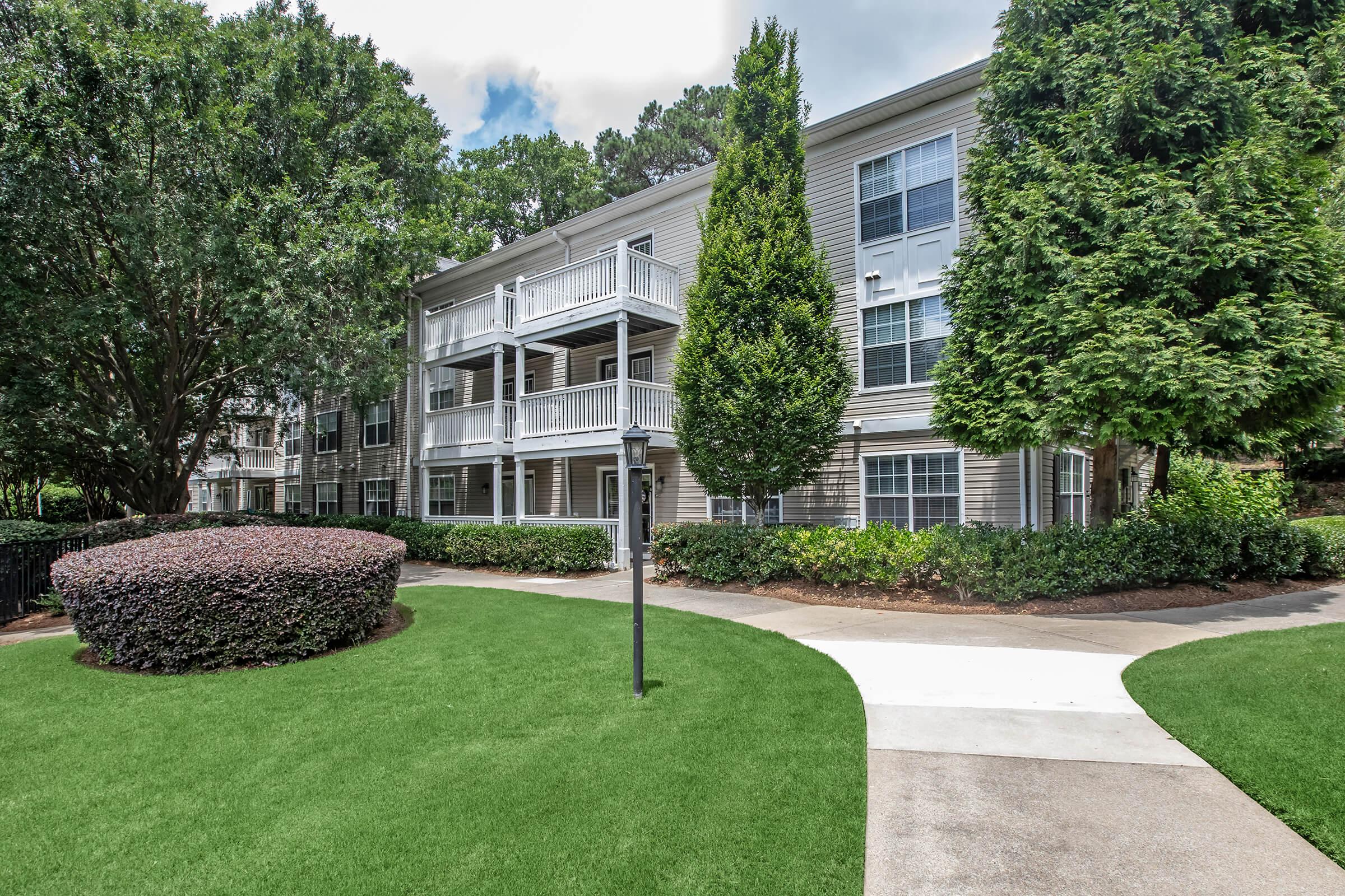 A well-maintained apartment building surrounded by lush green lawns and neatly trimmed bushes. The building features balconies, suggesting a residential community. Pathways are lined with greenery, creating a welcoming atmosphere. The scene is bright and inviting, set against a clear sky.