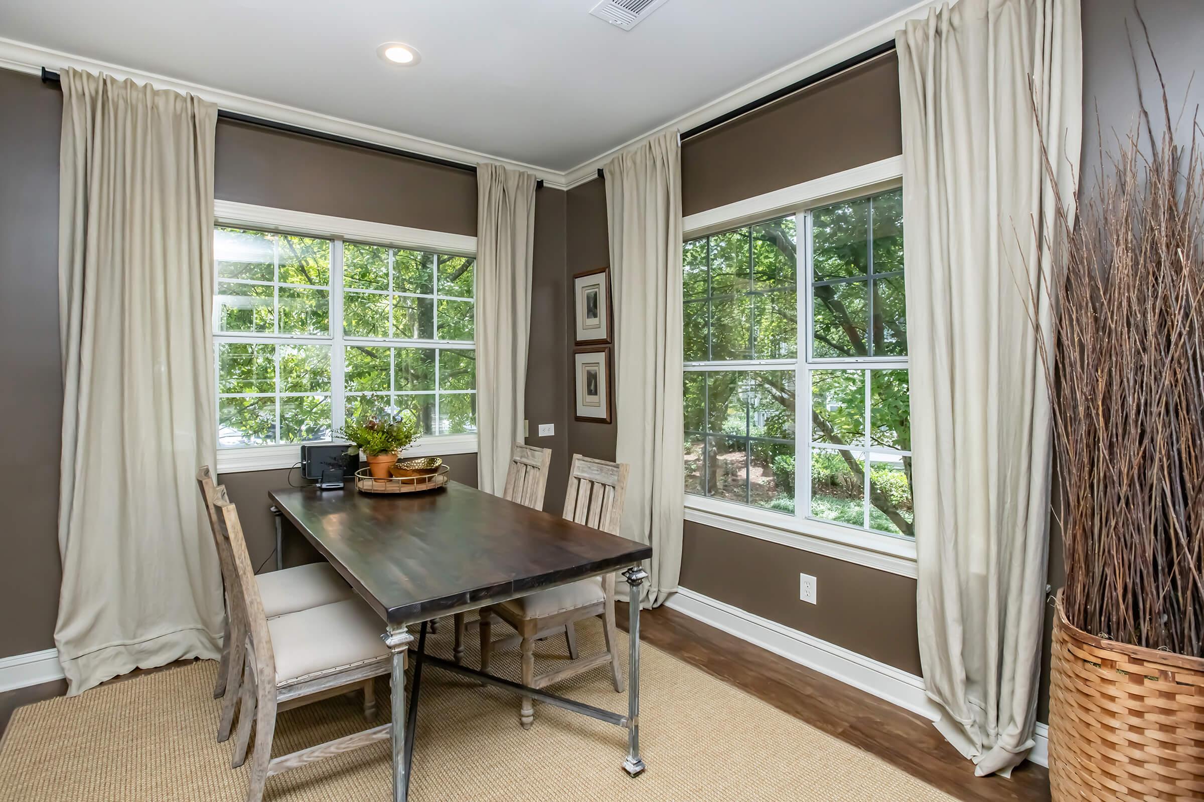 A cozy dining area featuring a dark wooden table surrounded by light-colored chairs. Large windows with cream curtains let in natural light, offering a view of greenery outside. A decorative arrangement sits in the center of the table, and a tall basket with branches adds texture to the room. The walls are painted brown for a warm ambiance.