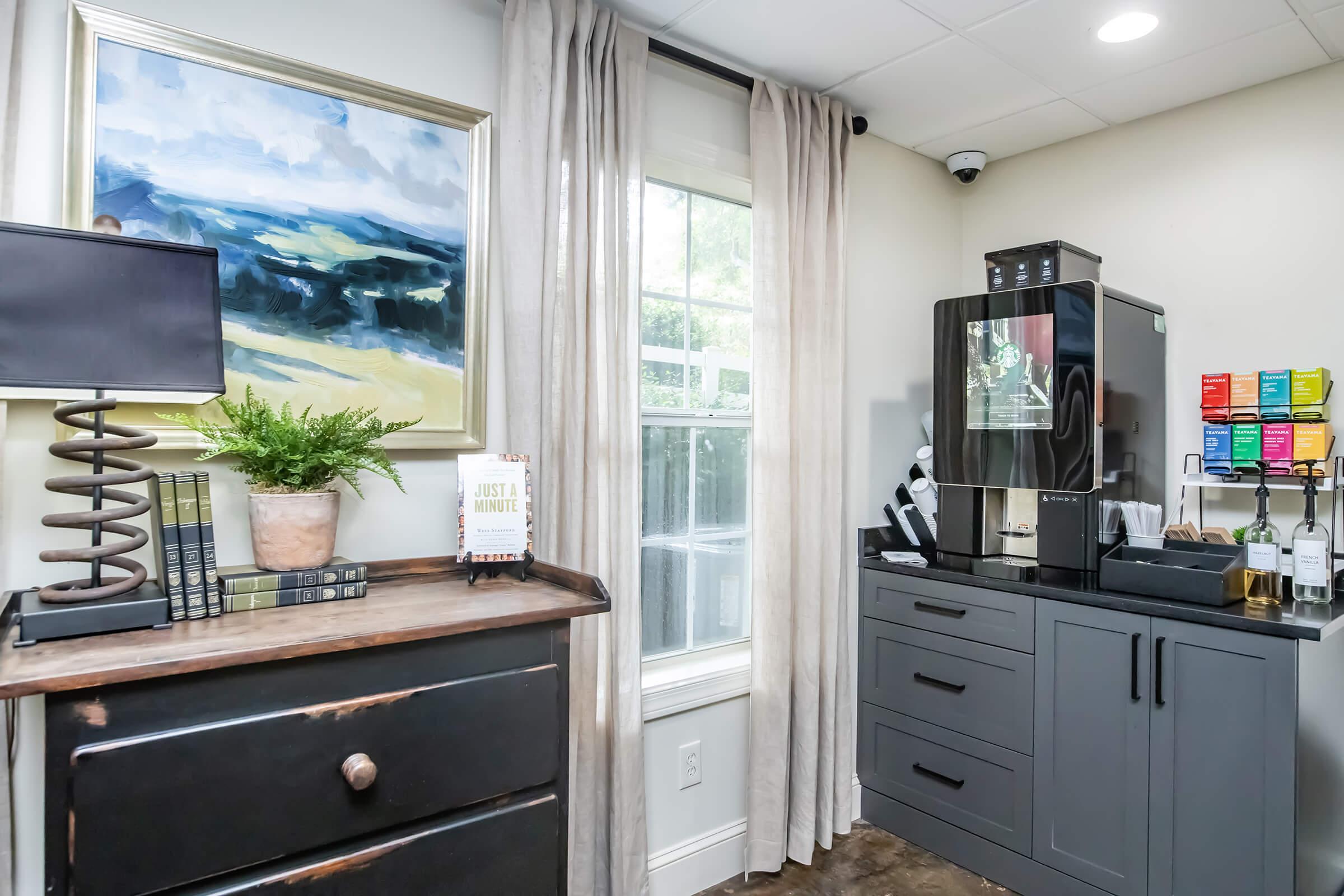 A stylish corner in a room featuring a dark wood dresser with books and a decorative plant, a modern black refrigerator with a color-coded water filtration system, and a table lamp. Natural light streams through a window with curtains, complemented by a landscape painting on the wall.