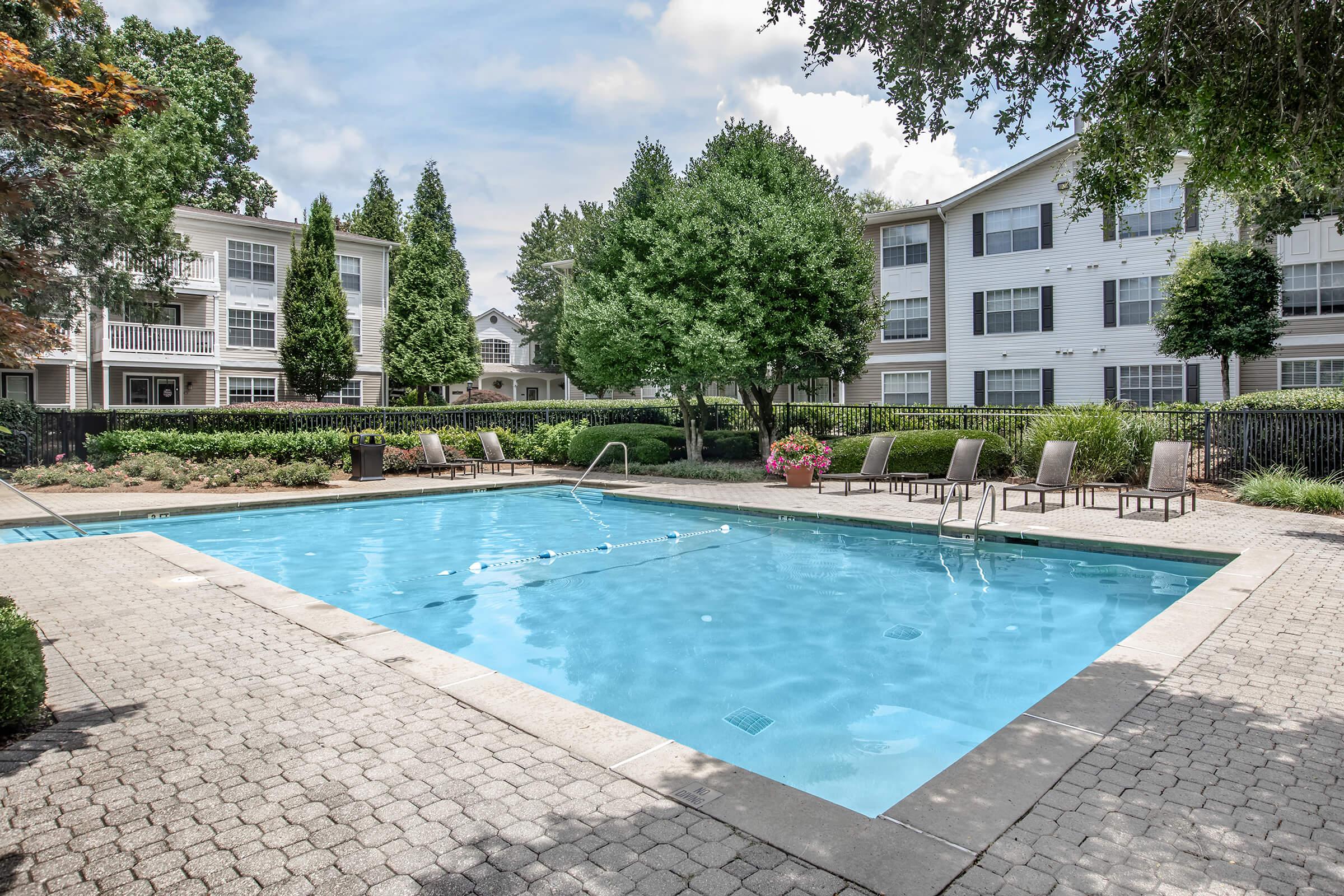 A clean outdoor swimming pool surrounded by lounge chairs, with manicured landscaping and trees in the background. The area features light gray stone paving, and there are nearby apartment buildings visible. The sky is partly cloudy, creating a pleasant and inviting atmosphere.