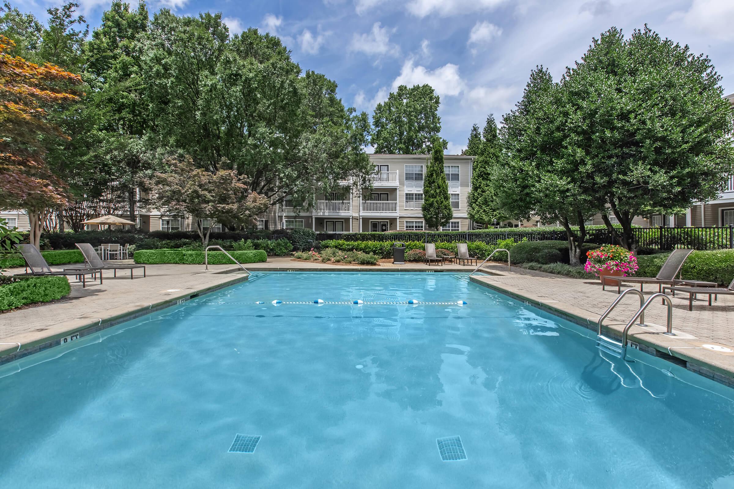 A tranquil swimming pool surrounded by lush greenery and landscaped gardens. Comfortable lounge chairs are positioned around the pool area. In the background, a multi-story building is partially visible, framed by trees under a clear blue sky with fluffy clouds.