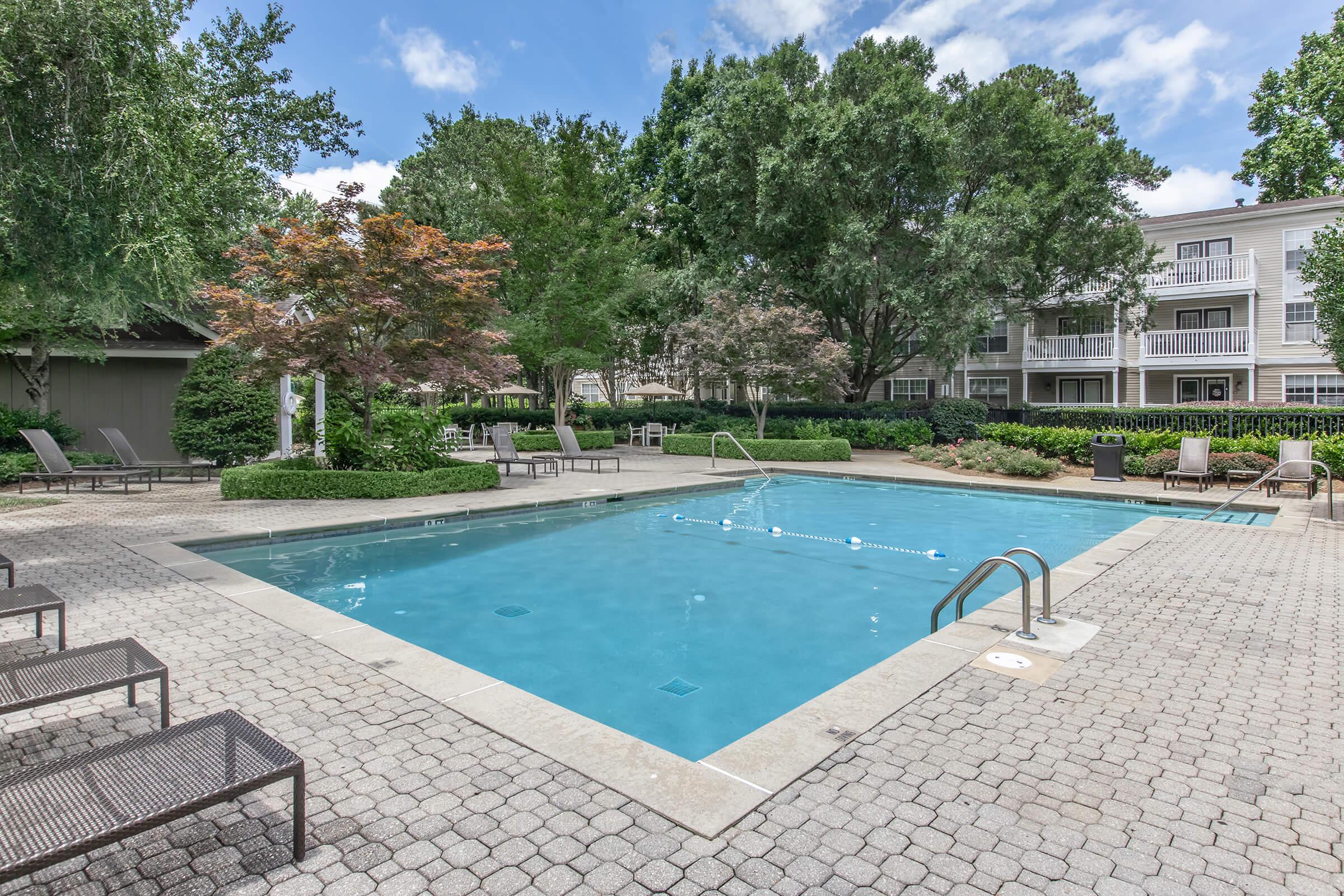 A swimming pool surrounded by a stone patio, lounge chairs, and trees. The pool features clear blue water with steps leading in, and is situated in a landscaped area with greenery and a few trees. In the background, there are residential buildings with balconies. The sky is partly cloudy.