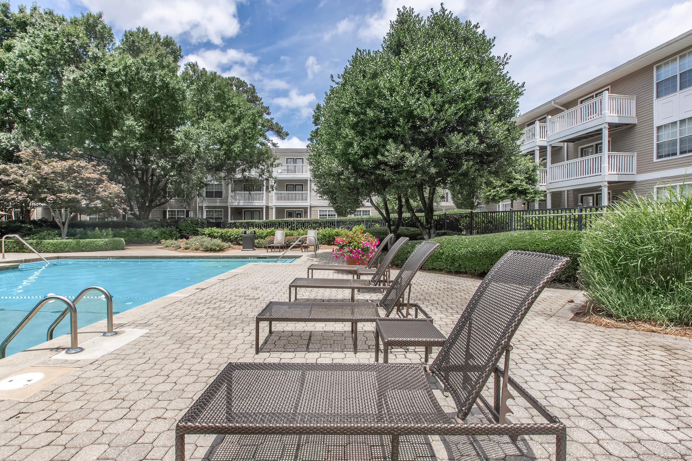 A tranquil pool area featuring several lounge chairs arranged alongside a clear blue swimming pool. Lush greenery and flowering plants surround the pool, with two residential buildings visible in the background under a partly cloudy sky. The scene conveys a relaxing outdoor atmosphere.