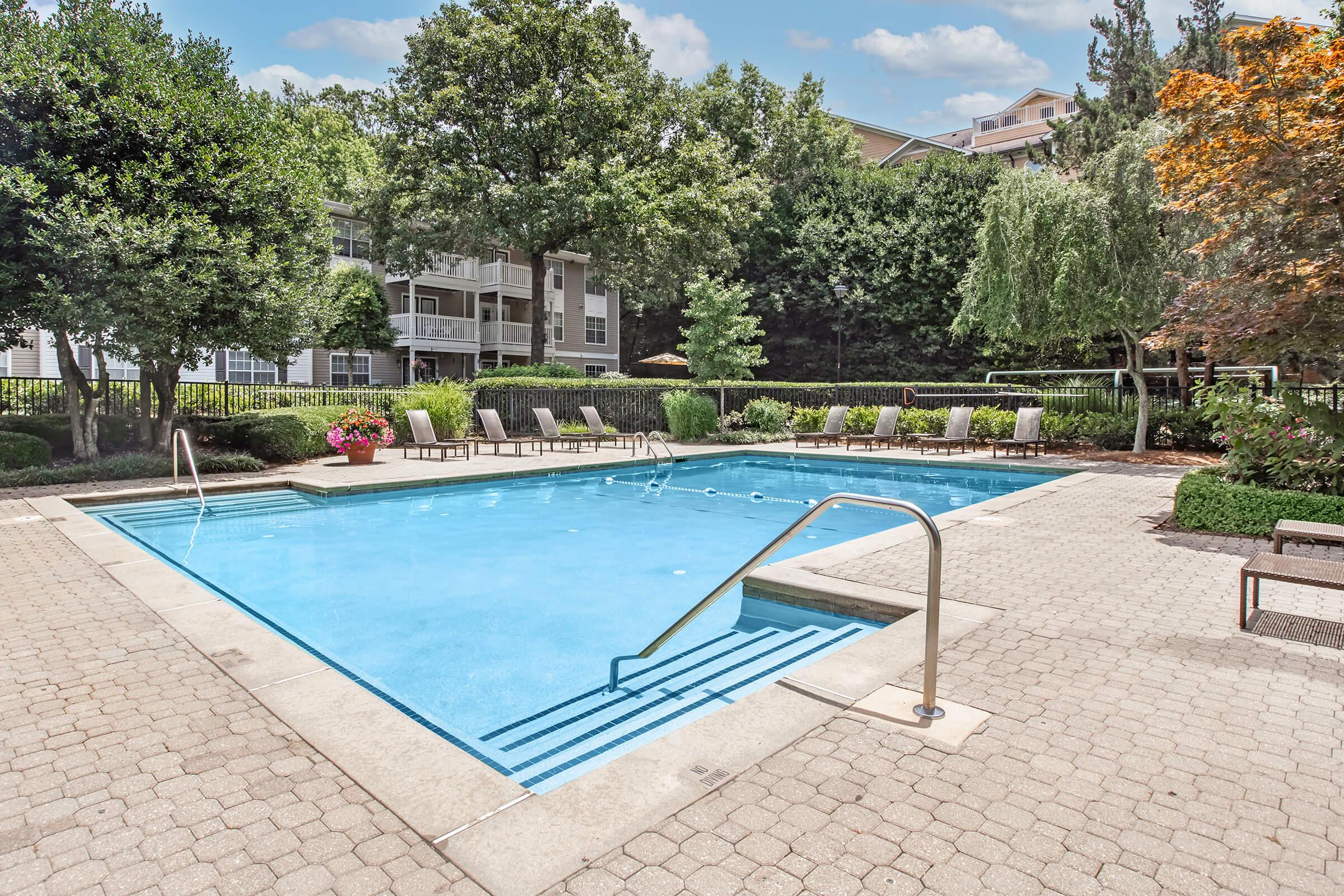 A clear swimming pool surrounded by a stone patio and lounge chairs. Lush greenery and flowering plants are visible in the background, along with apartment buildings. The sky is partly cloudy, creating a bright and inviting atmosphere.