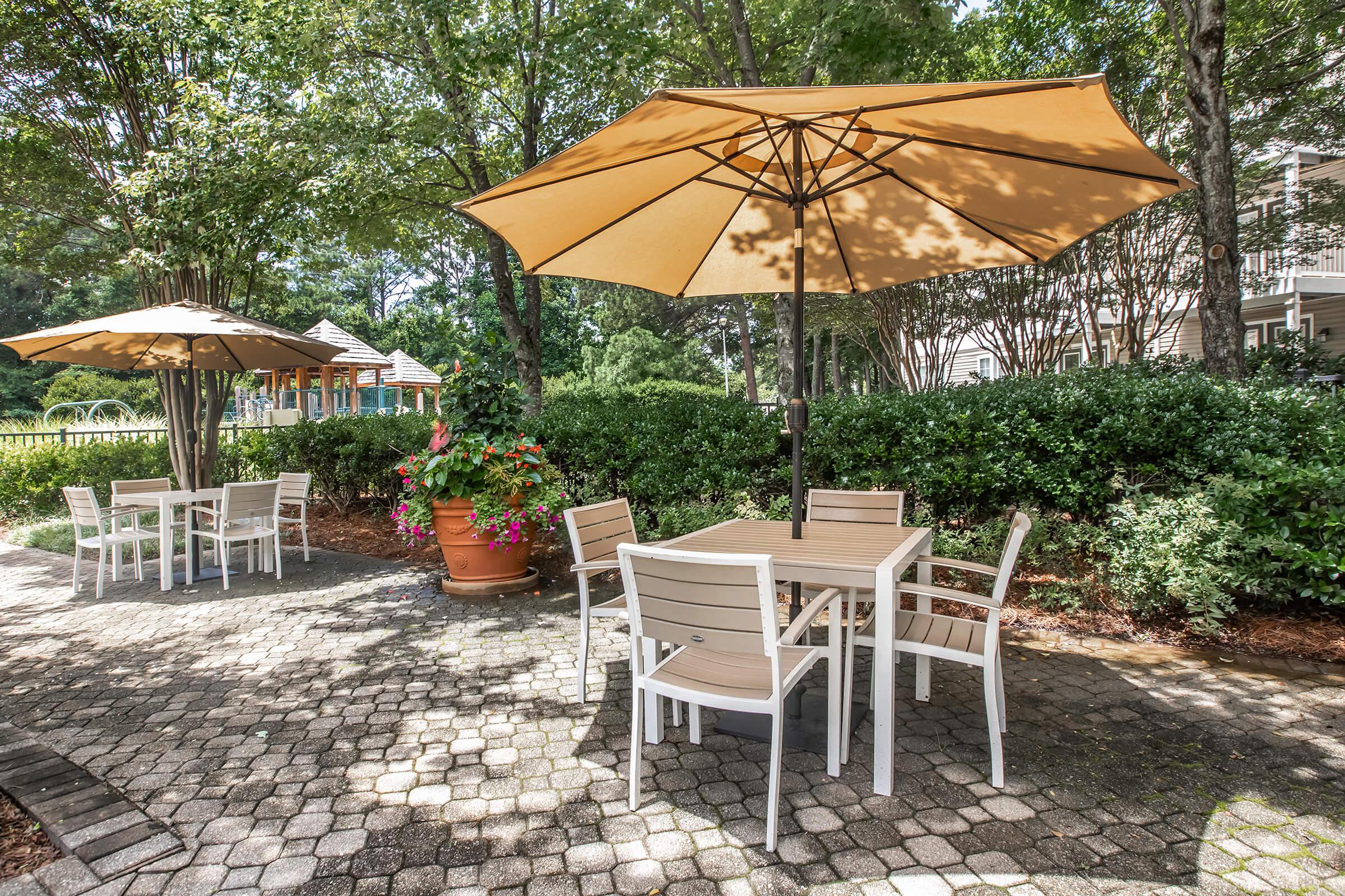 Outdoor seating area featuring wooden tables and white chairs under large beige umbrellas. Surrounded by greenery and flower pots, the setting is peaceful and inviting, perfect for relaxation. Pathways made of cobblestones enhance the tranquil ambiance.