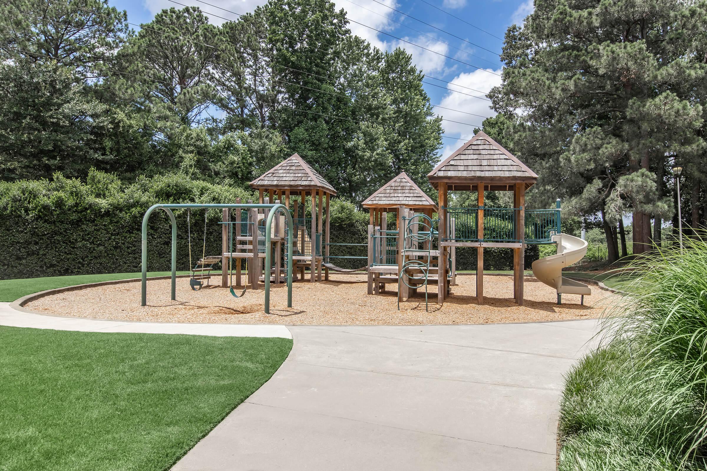 A playground featuring two wooden play structures with roofs, surrounded by green grass and trees. One structure includes a slide and climbing features, located on a sandy area with a concrete pathway leading to it. The sky is partly cloudy, creating a bright, inviting atmosphere.
