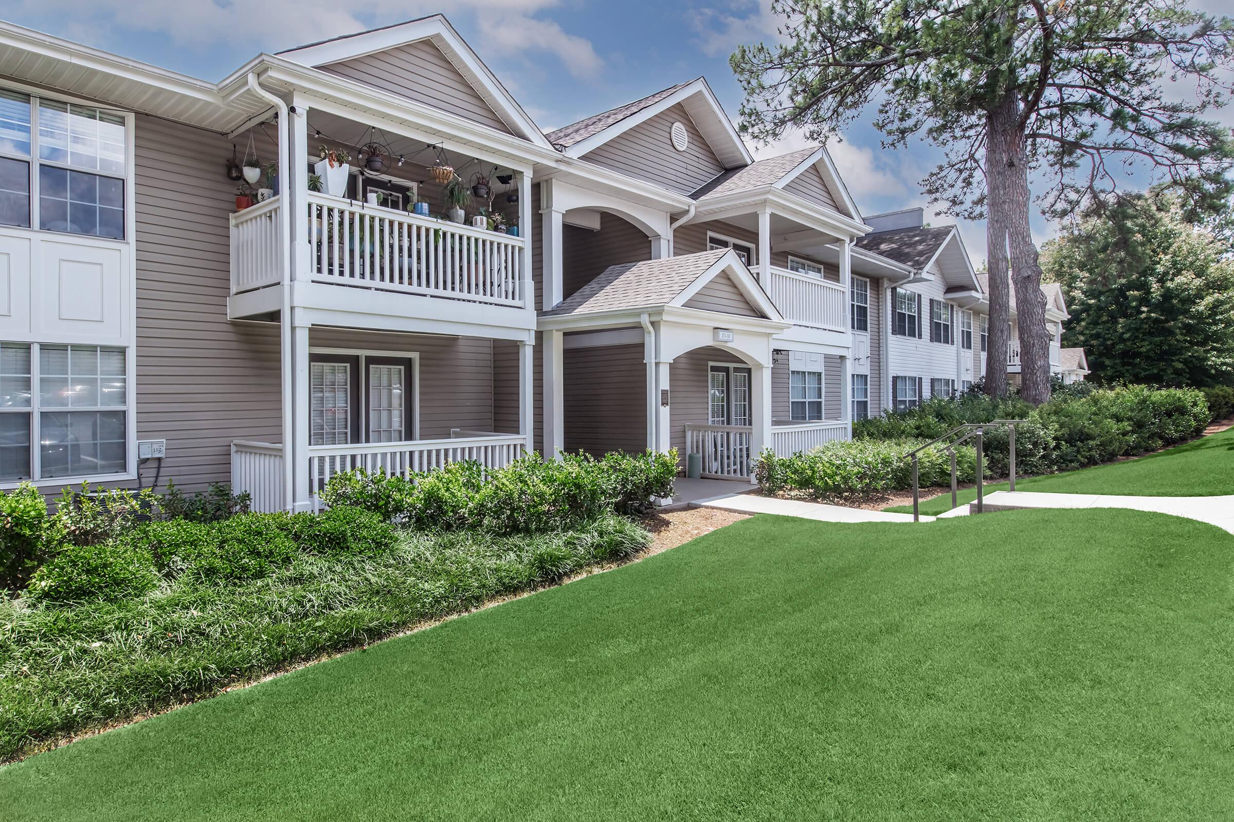 A view of a modern apartment complex featuring multiple units with balconies. The exterior has light-colored siding and well-maintained landscaping with lush green grass and shrubs. The setting is bright with a clear sky, emphasizing a clean and inviting residential atmosphere.
