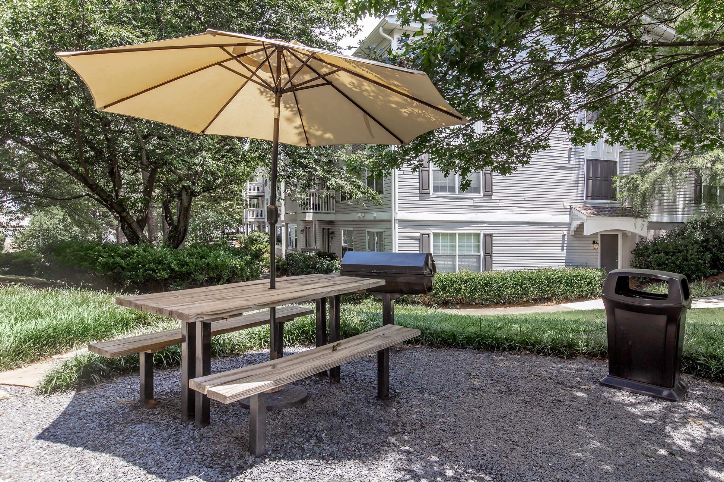 A picnic table with benches is shaded by a large umbrella in a landscaped area. Nearby, there is a grill and a trash can, with a background of apartment buildings and greenery. The scene is inviting for outdoor gatherings or meals.