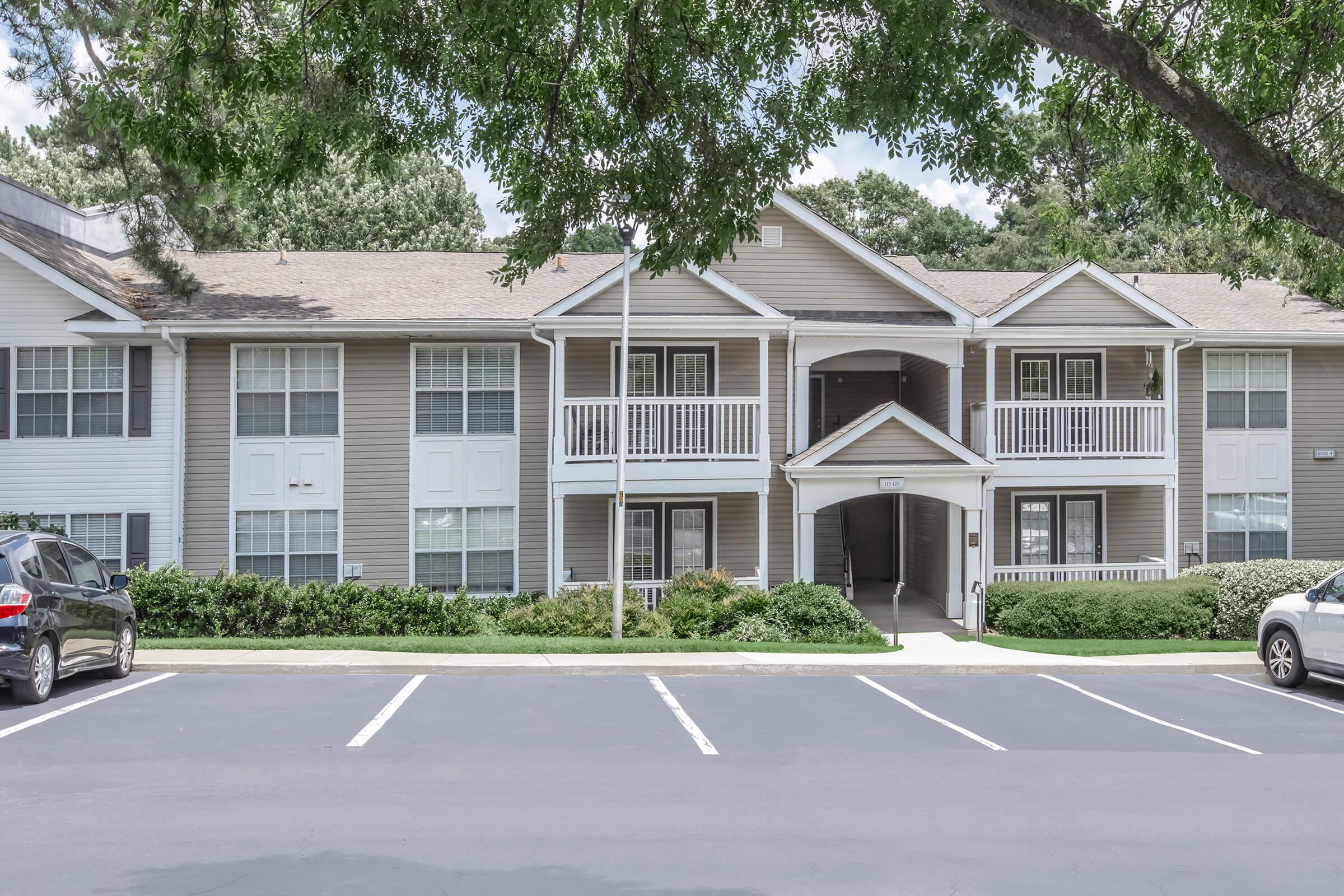 A three-story residential apartment building with a blend of light and dark siding. The entrance features a covered porch with white railings. The surrounding area includes greenery and a paved parking lot with several parked cars. Trees provide shade in the foreground.