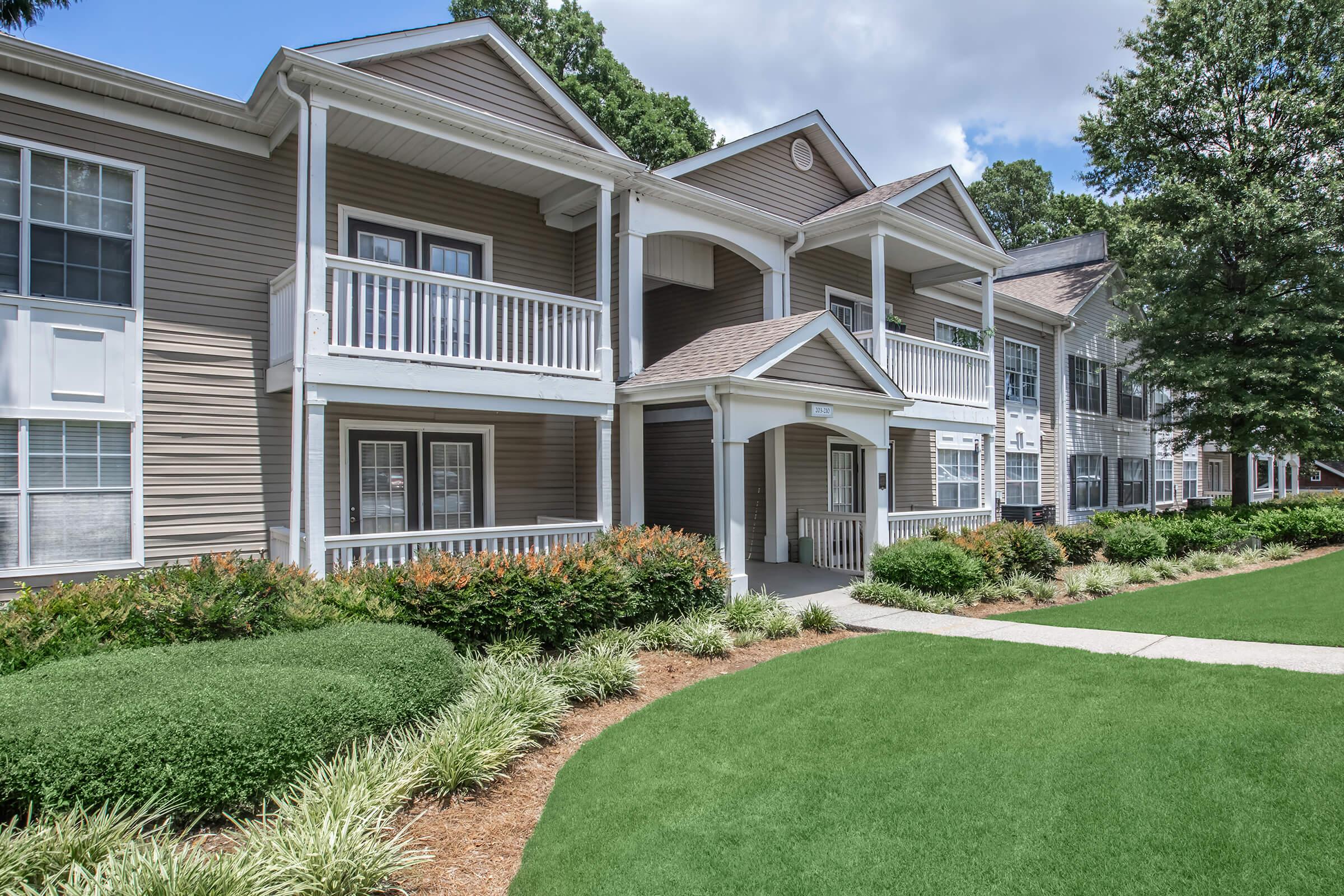 A well-maintained apartment complex with a light brown exterior, featuring multiple balconies, surrounded by neatly trimmed bushes and vibrant greenery. A walking path leads through the landscaped yard, enhancing the inviting atmosphere of the residence.