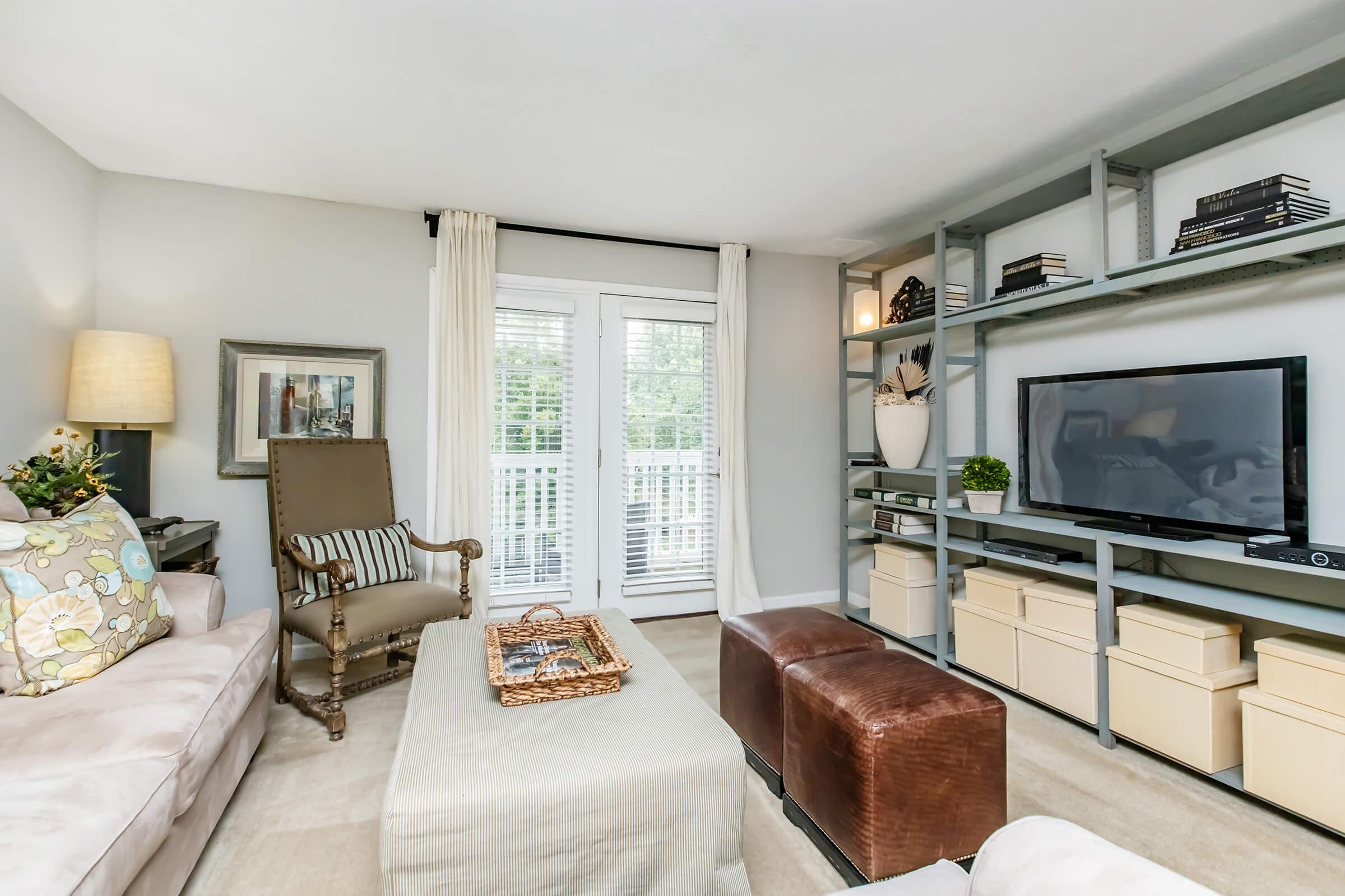 A cozy living room featuring a light-colored sofa, an armchair, and a coffee table. There is a TV on a shelving unit with books and decor, and light curtains covering a patio door. A warm ambience is created with soft lighting and neutral tones, complemented by a touch of greenery.