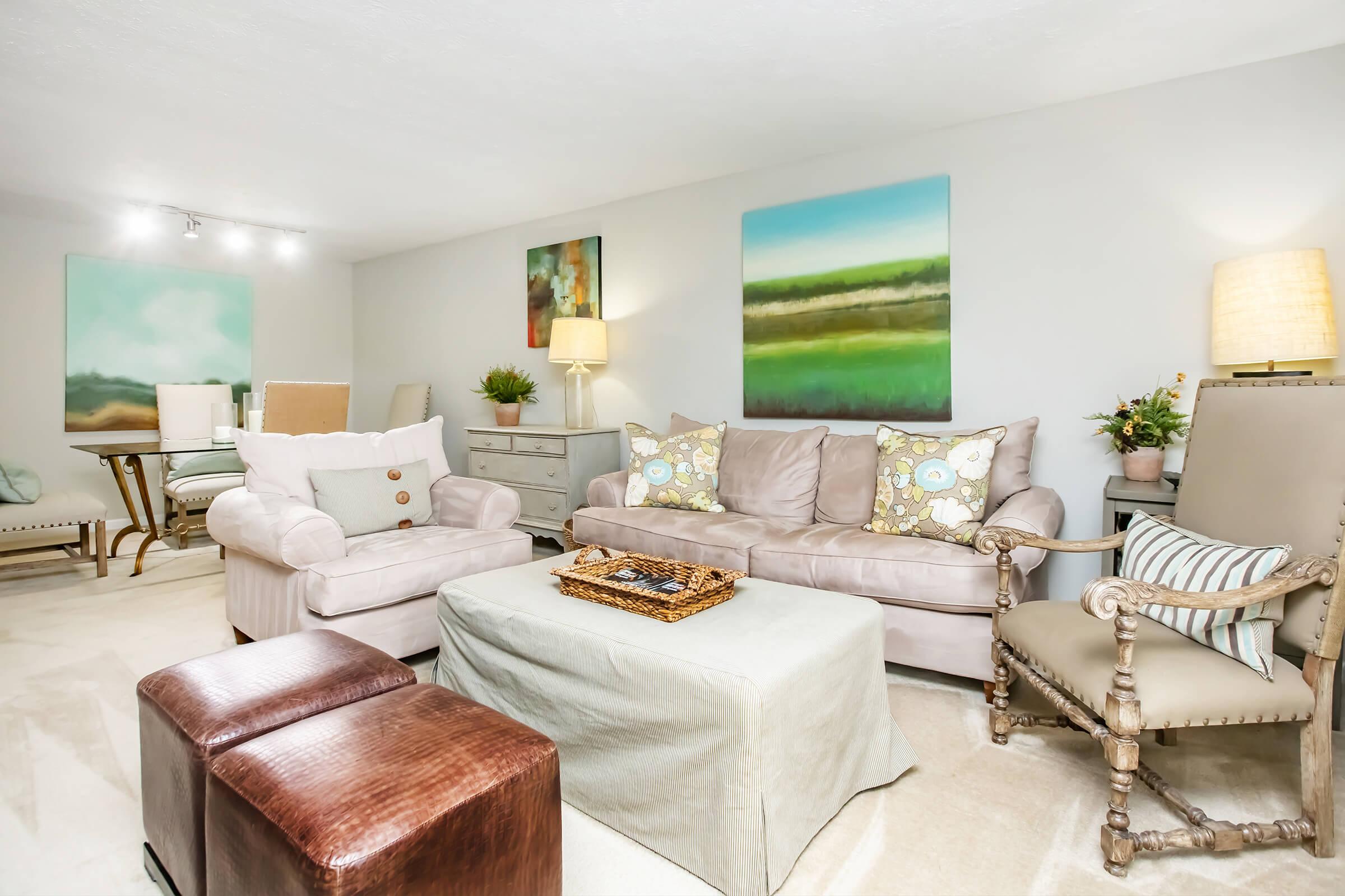 A cozy living room featuring a light-colored sofa with decorative pillows, a beige armchair, a wooden coffee table with a tray, and two brown ottomans. The walls are adorned with abstract paintings in soft colors, and the space is well-lit with lamps and natural light. A touch of greenery adds warmth.