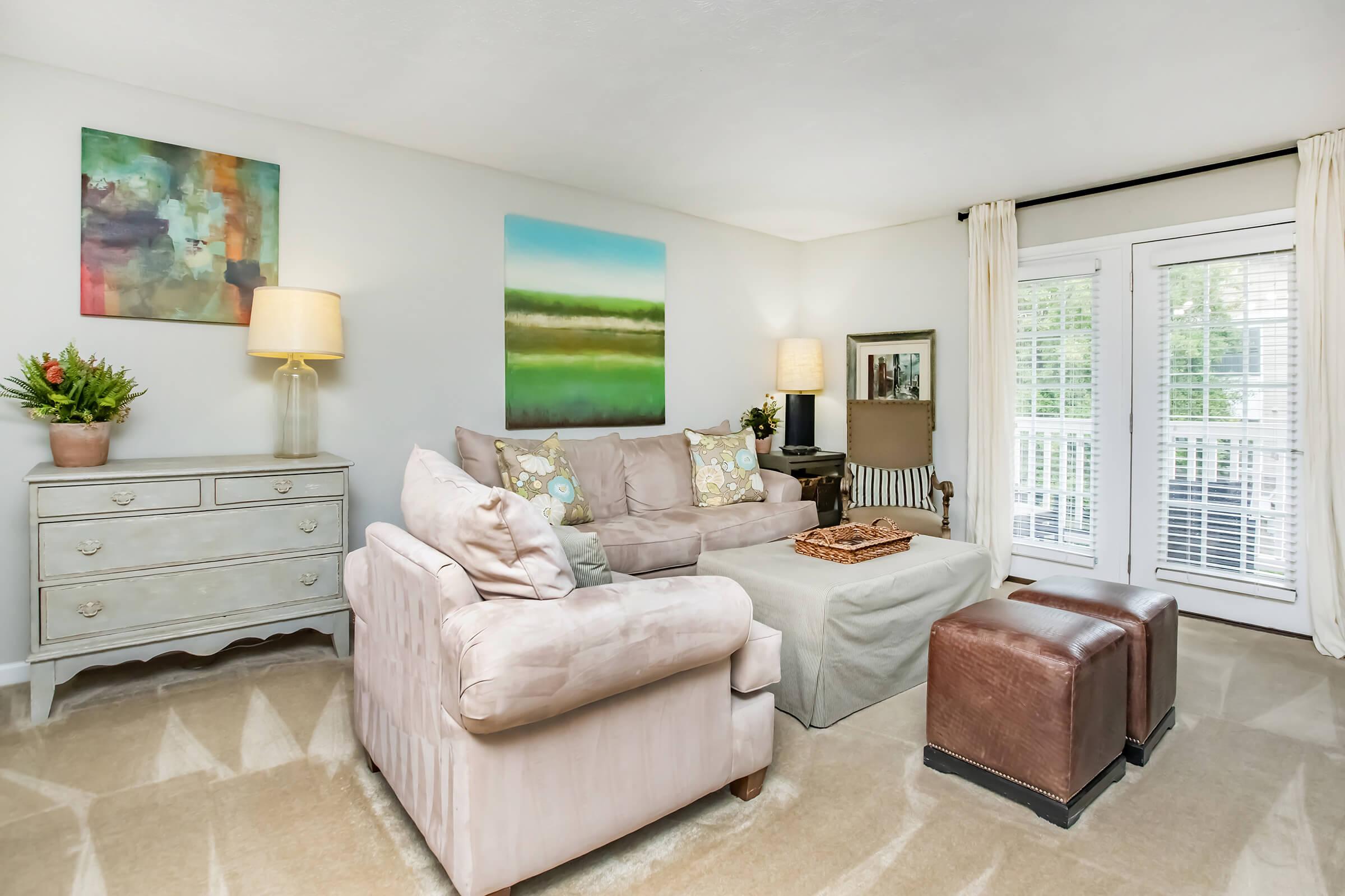 A cozy living room featuring a light-colored sofa with decorative pillows, a wooden coffee table, and two leather ottomans. There is a dresser adorned with a lamp and plants, along with colorful artwork on the walls. Natural light streams in through white plantation shutters, enhancing the inviting atmosphere.
