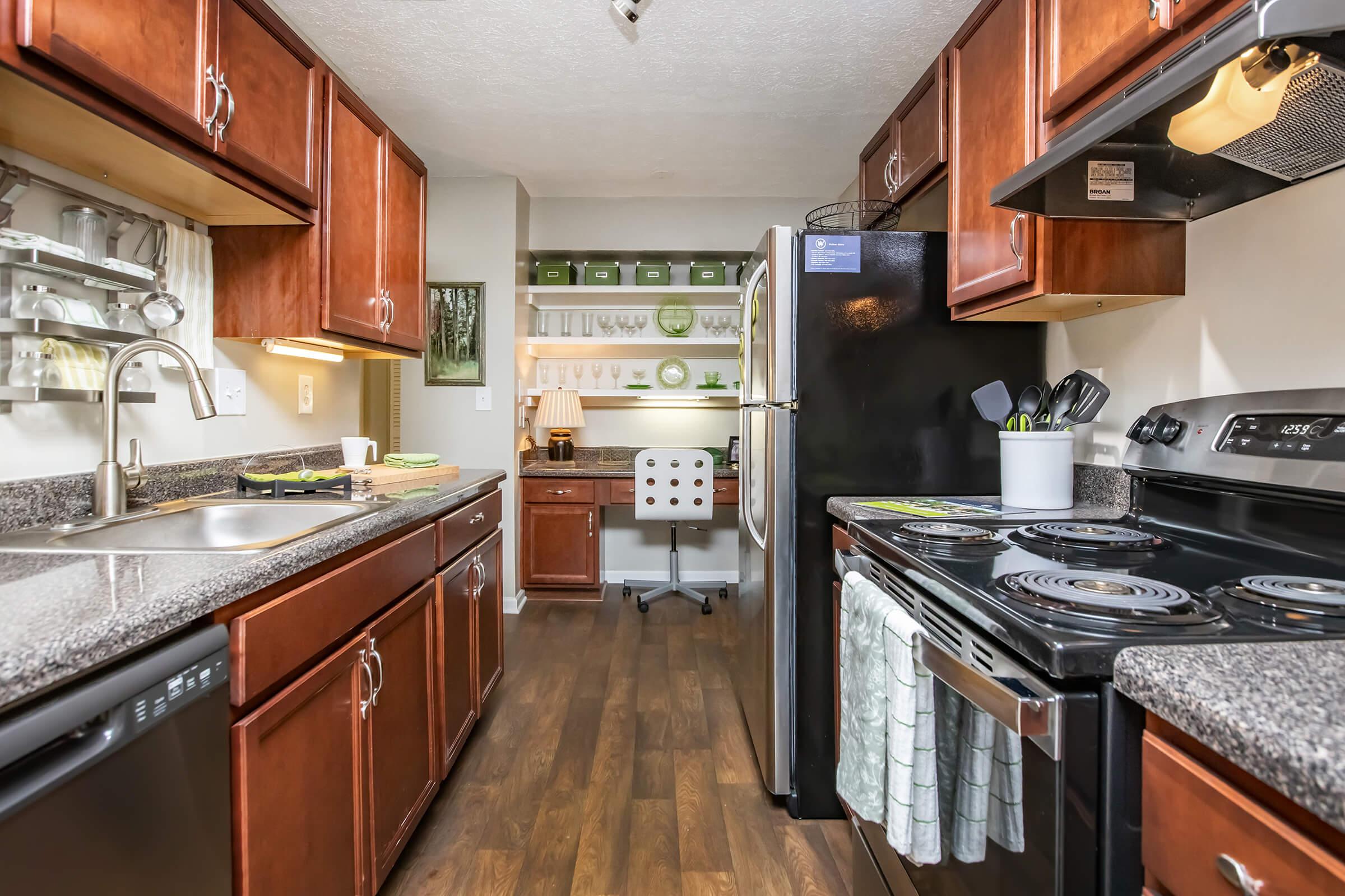 A modern kitchen featuring dark wooden cabinets, granite countertops, and stainless steel appliances. The layout includes a black refrigerator, an oven, and a sink with a tap. A small workspace with a white chair is visible, along with shelves displaying green decorative items. The floor is dark wood.