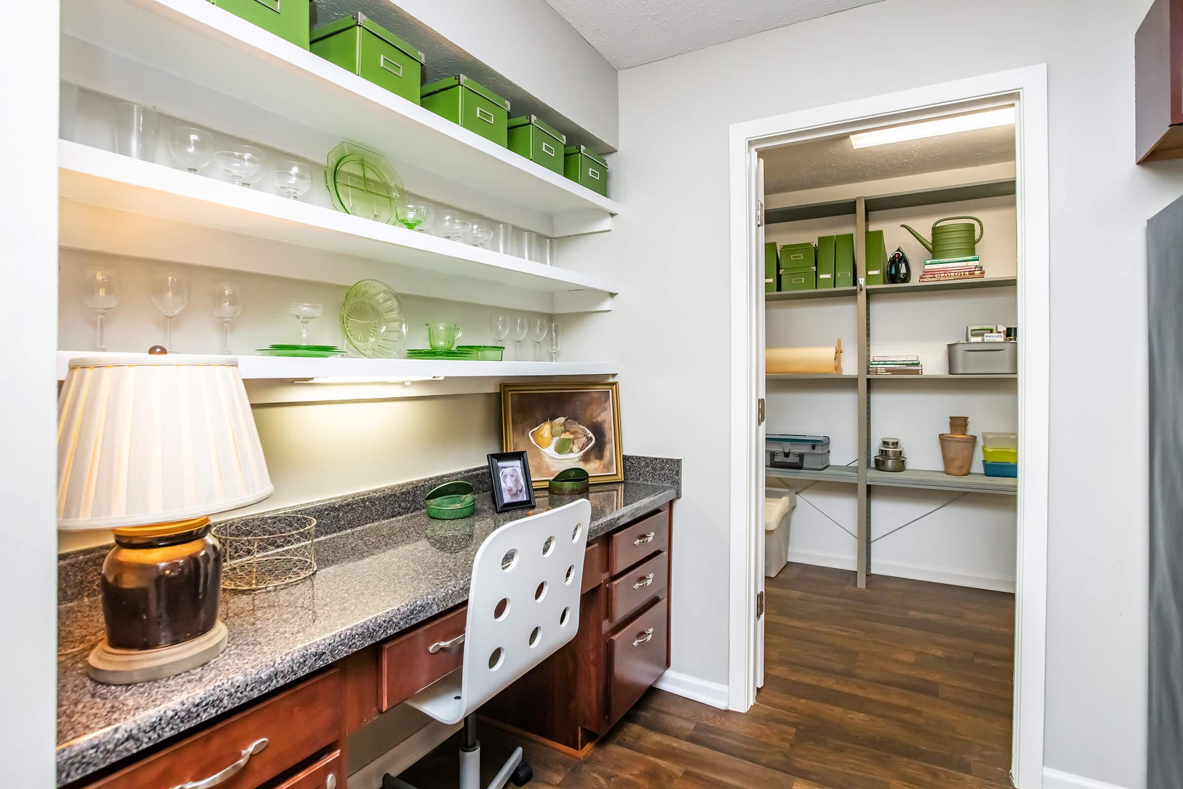 A neatly organized pantry or workspace featuring a countertop with a lamp, photo frame, and decorative items. Shelves above display glassware and green storage boxes. A door leads to a second storage space, which is also organized with shelves and various items. The flooring is wooden, adding warmth to the space.