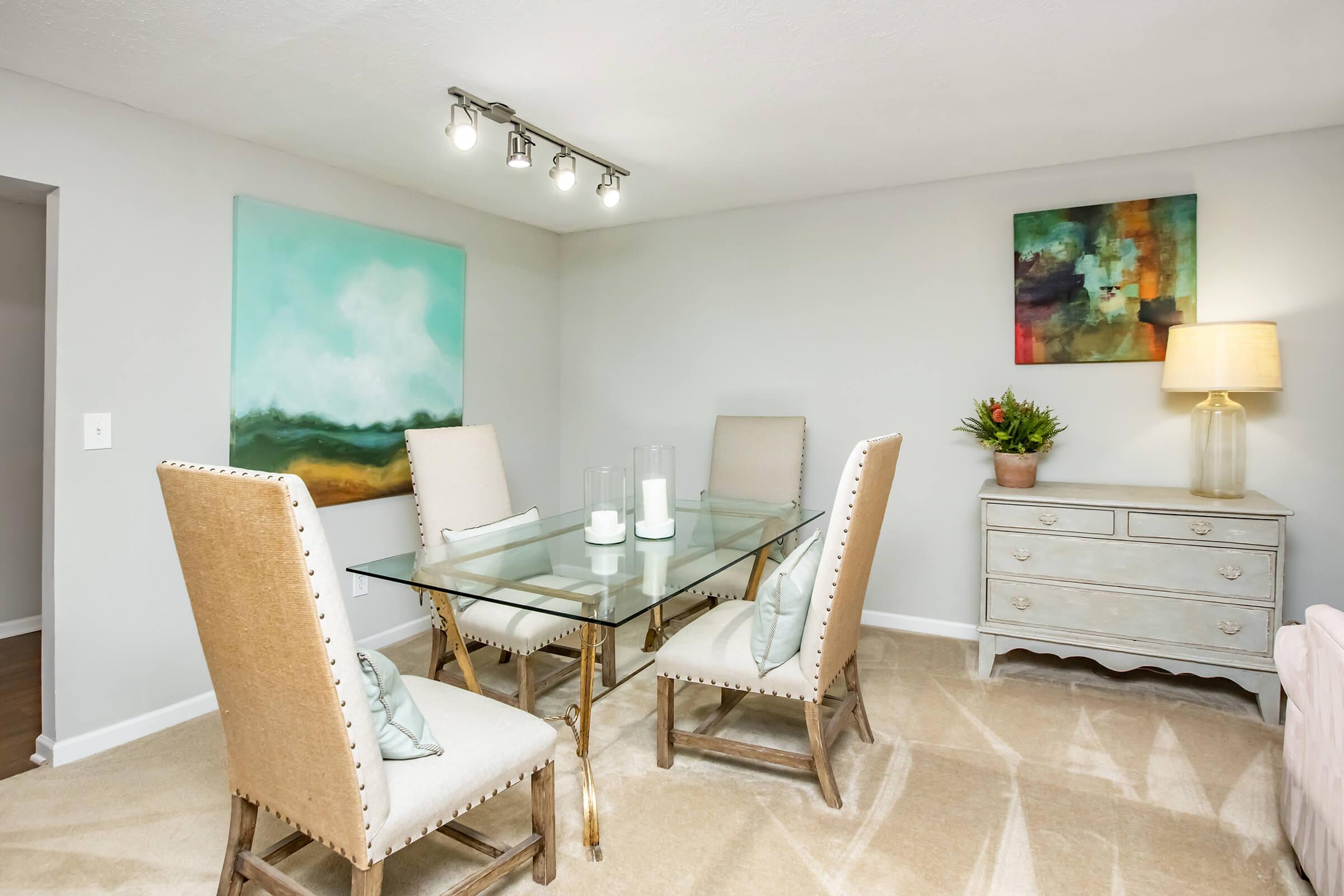 Modern dining area featuring a glass table surrounded by four elegant upholstered chairs. The room has light gray walls, with two abstract paintings and a stylish lamp beside a decorative chest. Natural light enhances the inviting atmosphere, complemented by a floral arrangement on the chest.