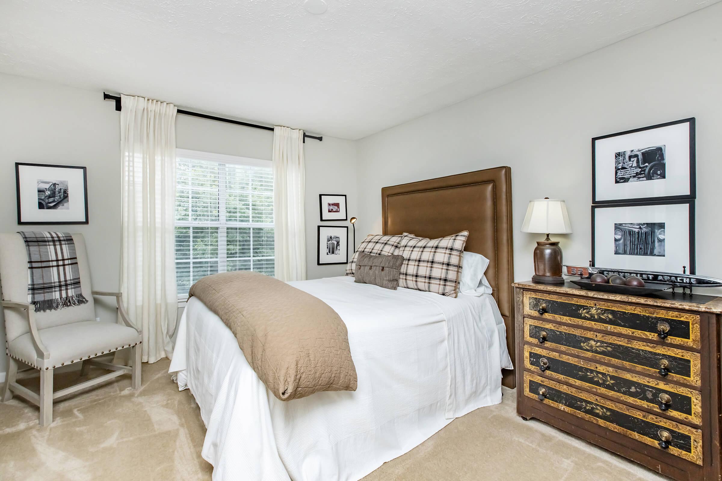 A cozy bedroom featuring a queen-sized bed with decorative pillows, a soft beige throw blanket, and a wooden nightstand with a lamp. A comfortable armchair is positioned nearby, and the walls are adorned with framed black-and-white photographs. Natural light filters through a window with sheer curtains.