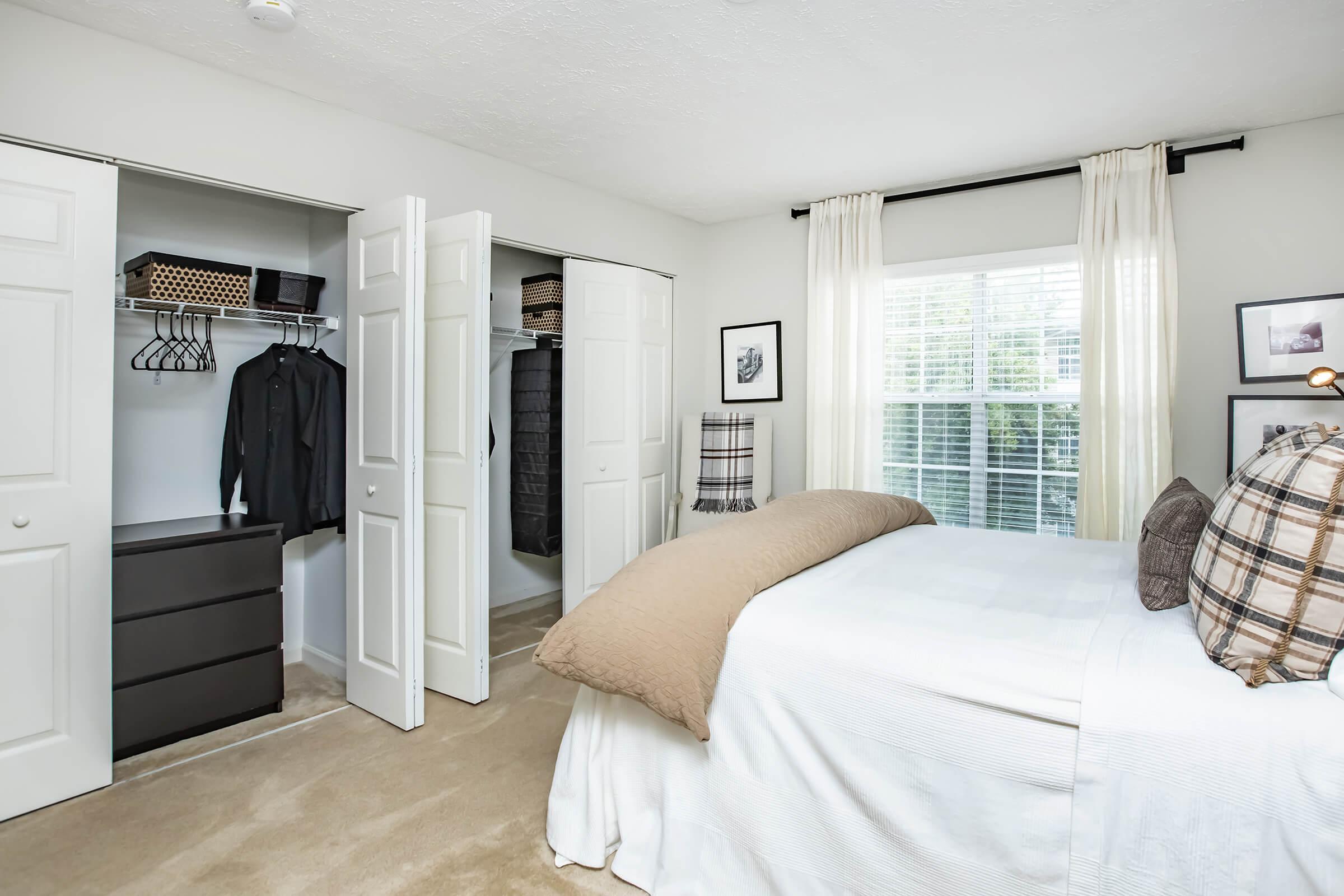 A cozy bedroom featuring a neatly made bed with a tan comforter, against a backdrop of light gray walls. Two closet doors are open, revealing organized clothing. A dresser is positioned beside the bed, and the room is well-lit with natural light coming through a window adorned with white curtains. Decorative wall art completes the space.