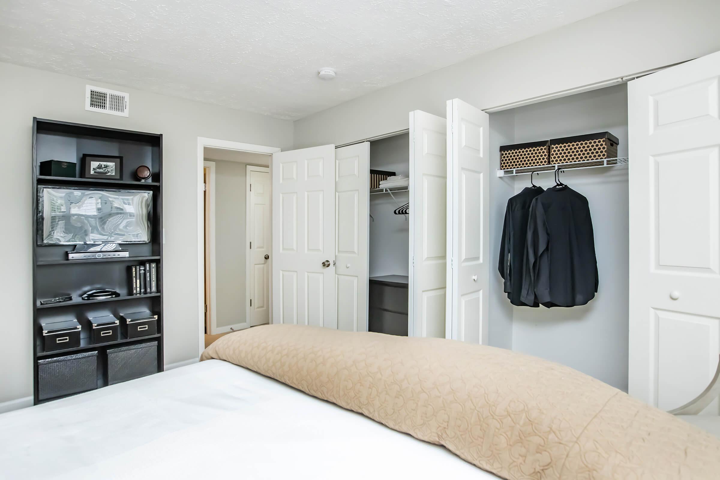 A well-lit bedroom featuring a neatly made bed with a light brown comforter. In the background, there are open closet doors showing hanging garments and shelves. A black bookshelf holds storage boxes and decorative items. The walls are a soft neutral color, creating a calm atmosphere.