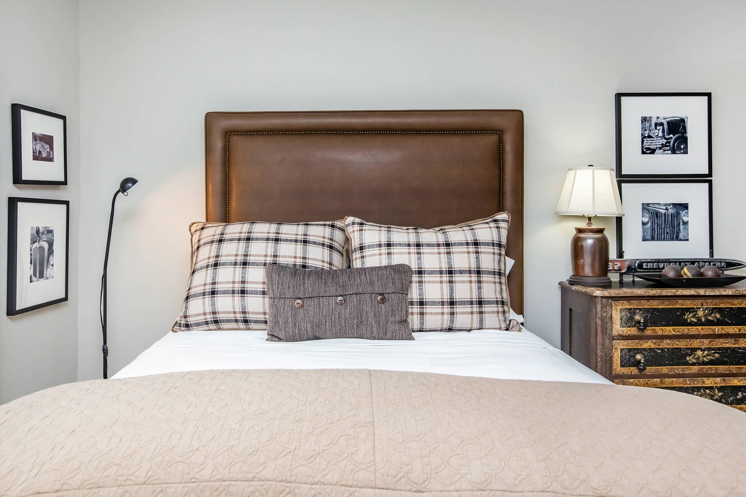 A cozy bedroom featuring a brown leather headboard with plaid pillows and a decorative cushion. Beside the bed, there's a small wooden nightstand with a lamp and framed black-and-white photos on the walls. The bedding is light-colored, creating a warm and inviting atmosphere.