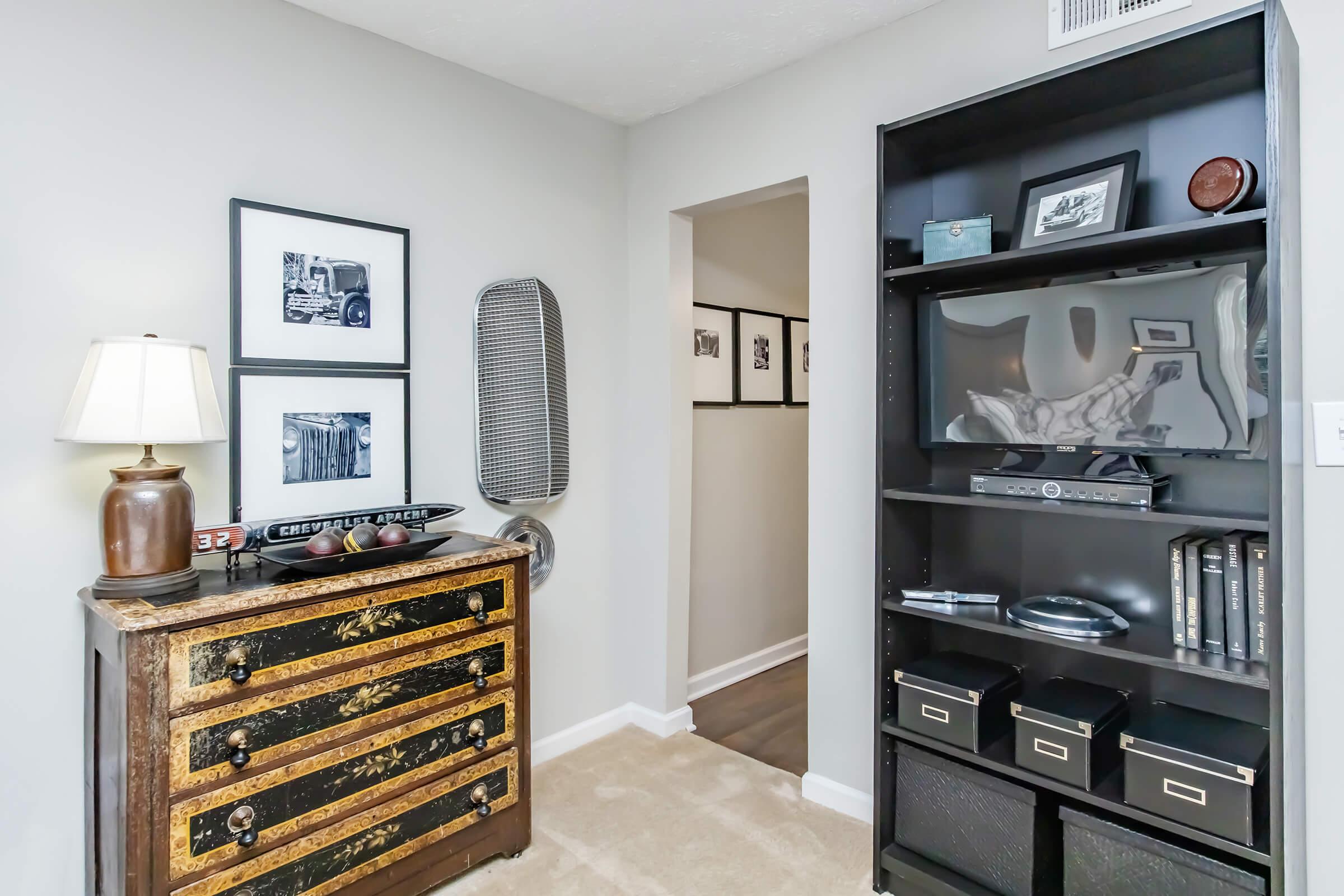 A cozy interior space featuring a vintage chest of drawers with decorative elements, a lamp, framed black-and-white photographs on the wall, a dark bookcase with various items, and a doorway leading to another room. The walls are painted gray, and the flooring appears to be a light-colored carpet.