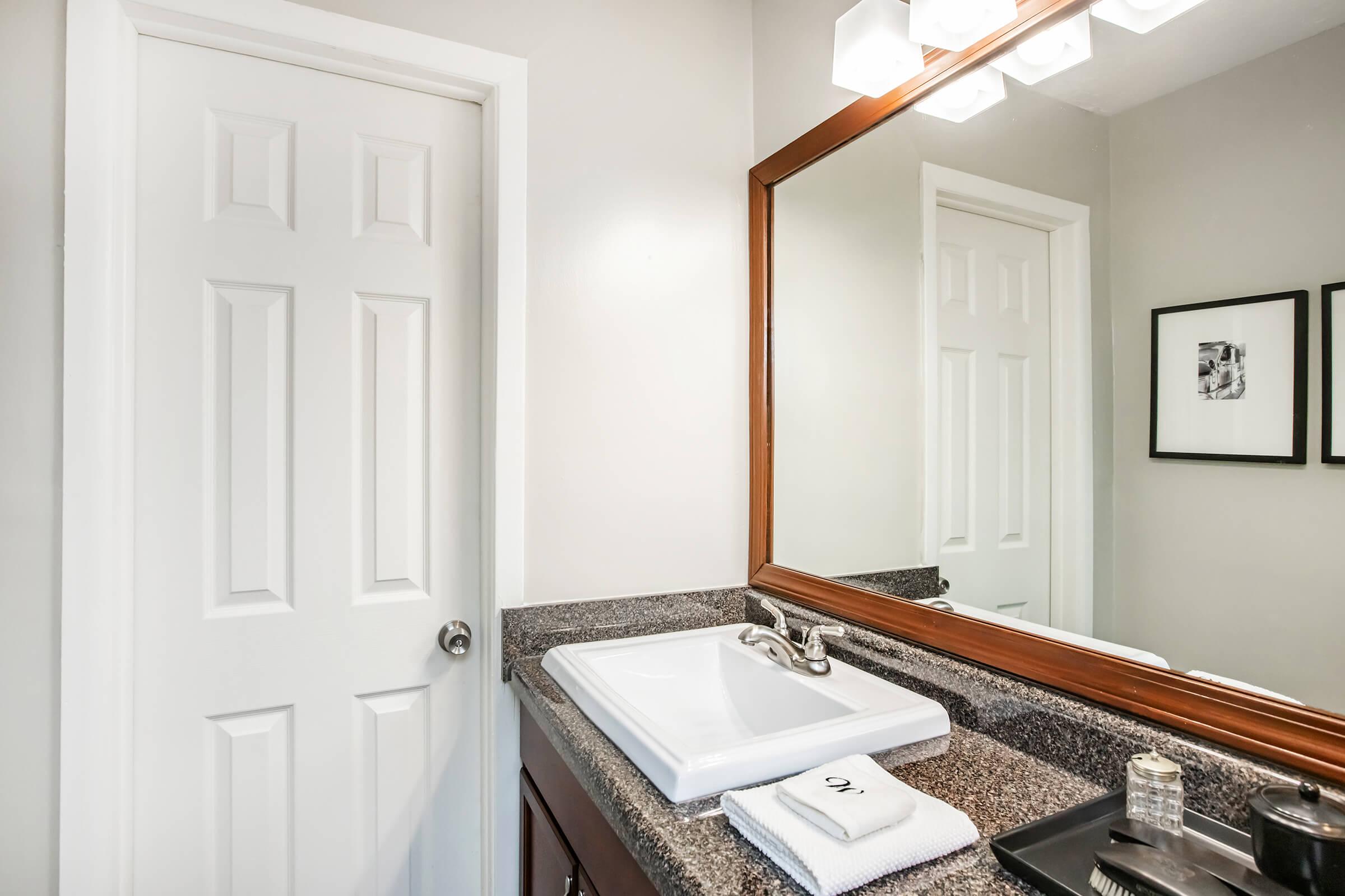 A modern bathroom featuring a white sink with a sleek faucet on a dark countertop. A large mirror with a wooden frame reflects the space. A white door leads out of the bathroom, and two framed black-and-white photos are mounted on the wall beside the mirror. Soft, neutral colors create a calming atmosphere.
