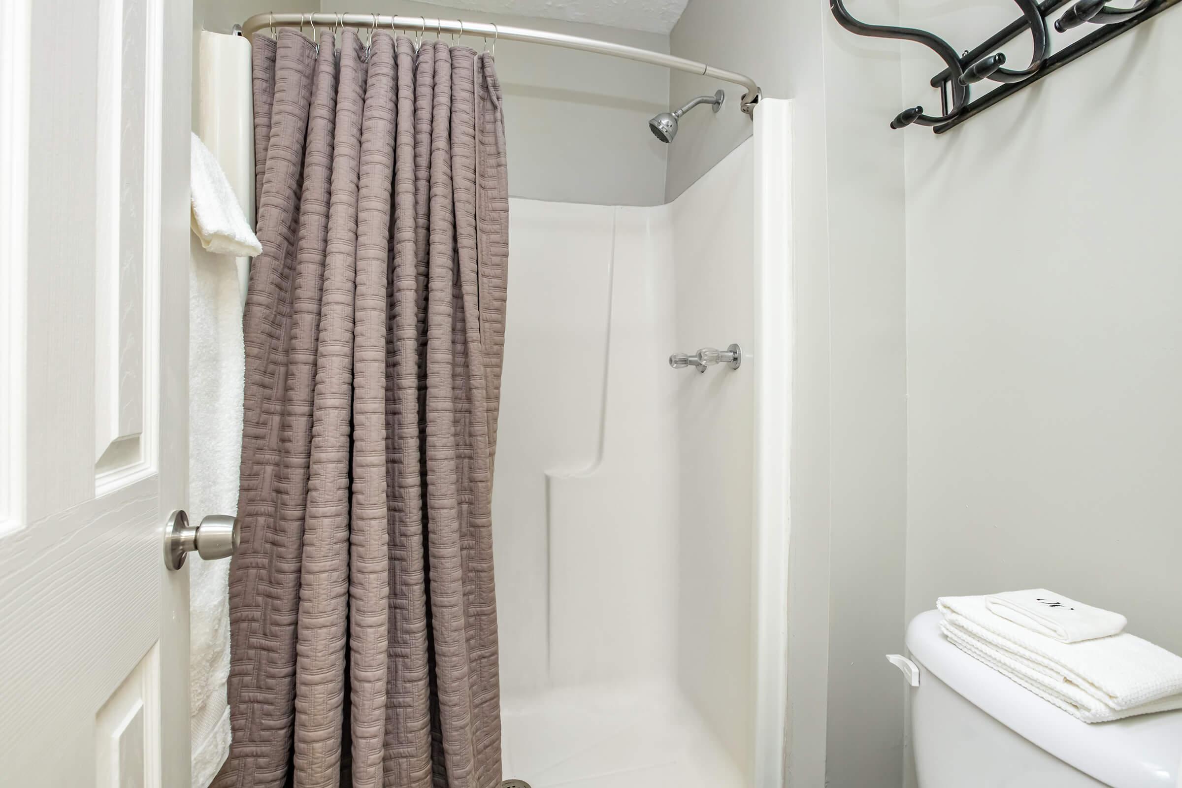 A small bathroom featuring a shower stall with a textured gray curtain, a white toilet beside it, and neatly folded white towels on the floor. The walls are painted in a light gray color, and there is a towel rack on the wall.