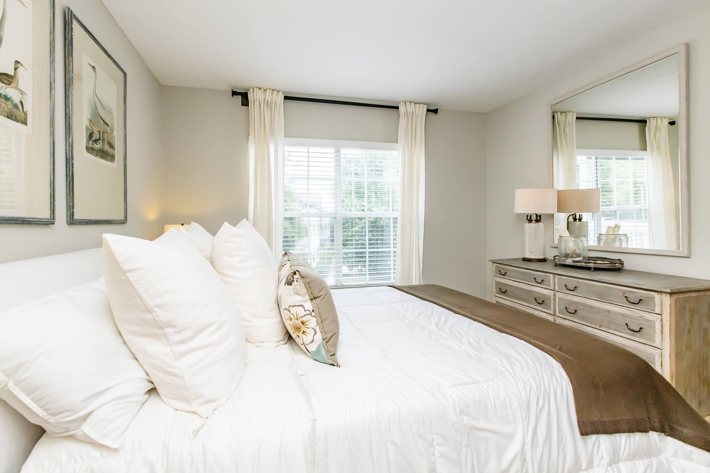 A cozy bedroom featuring a neatly made bed with fluffy pillows and a decorative throw. Light curtains frame a window that allows natural light to fill the room. A mirrored dresser and a lamp add to the elegant decor, creating a warm and inviting atmosphere.