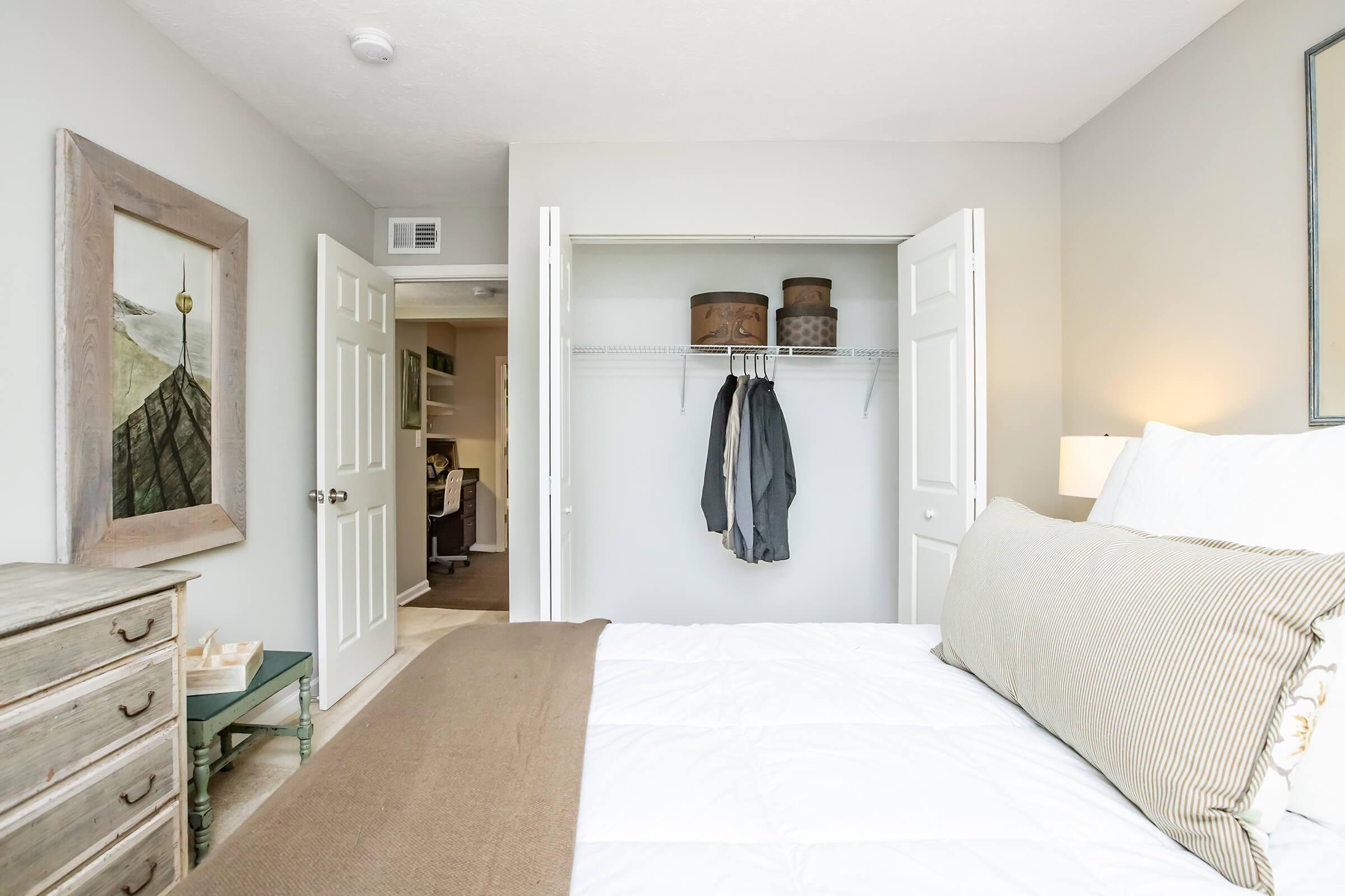 A cozy bedroom featuring a neatly made bed with white linens and decorative pillows. A closet with sliding doors is open, revealing hanging clothes and storage bins. A wooden dresser and artwork on the wall add warmth to the inviting space, complemented by soft lighting from a bedside lamp.