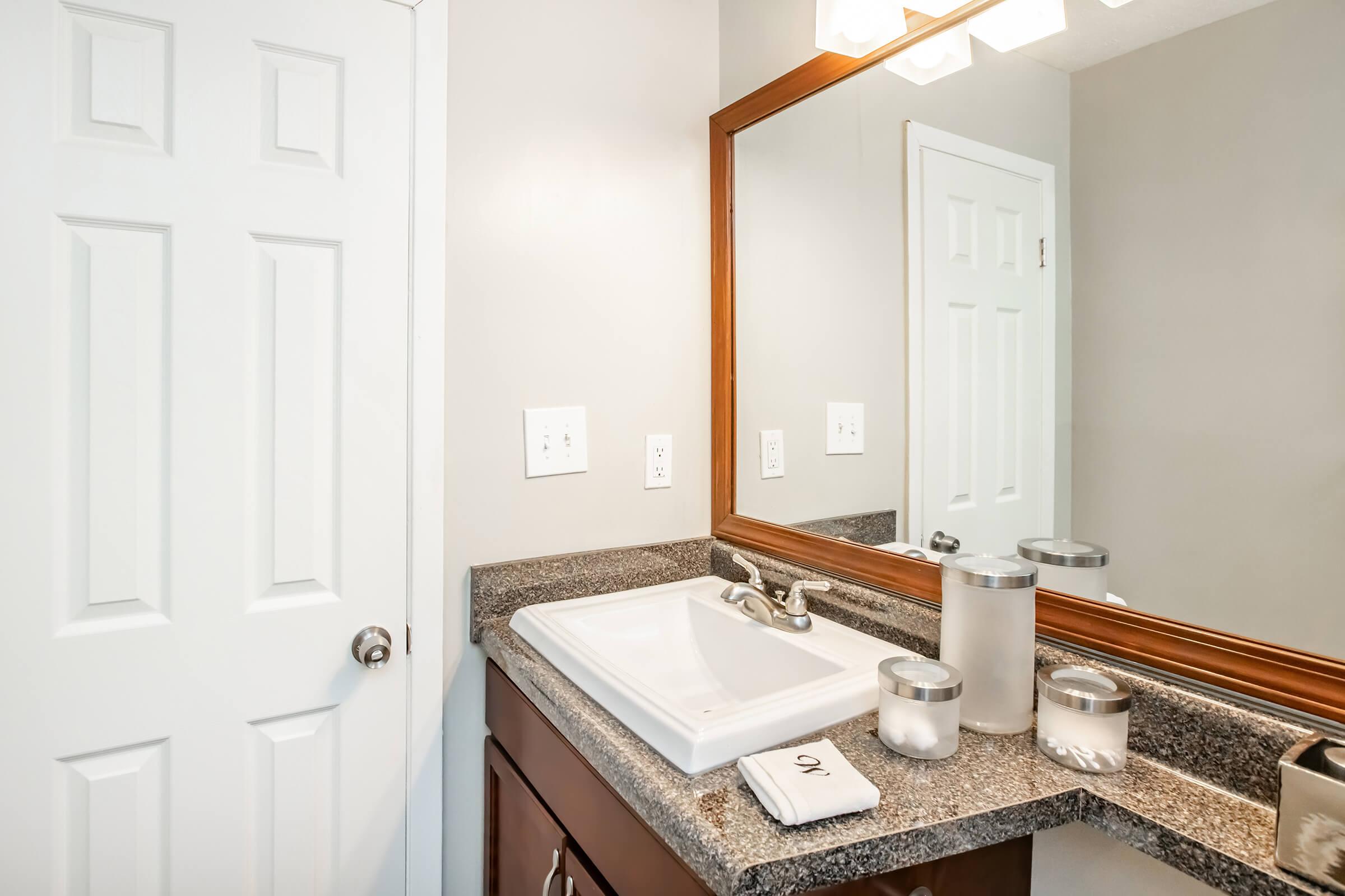 A bathroom countertop with a sink and mirror. The countertop features gray granite, a white sink, and several decorative items including glass containers. A white towel with the letter "O" is placed on the surface. A white door is visible in the background. The walls are painted a light gray.