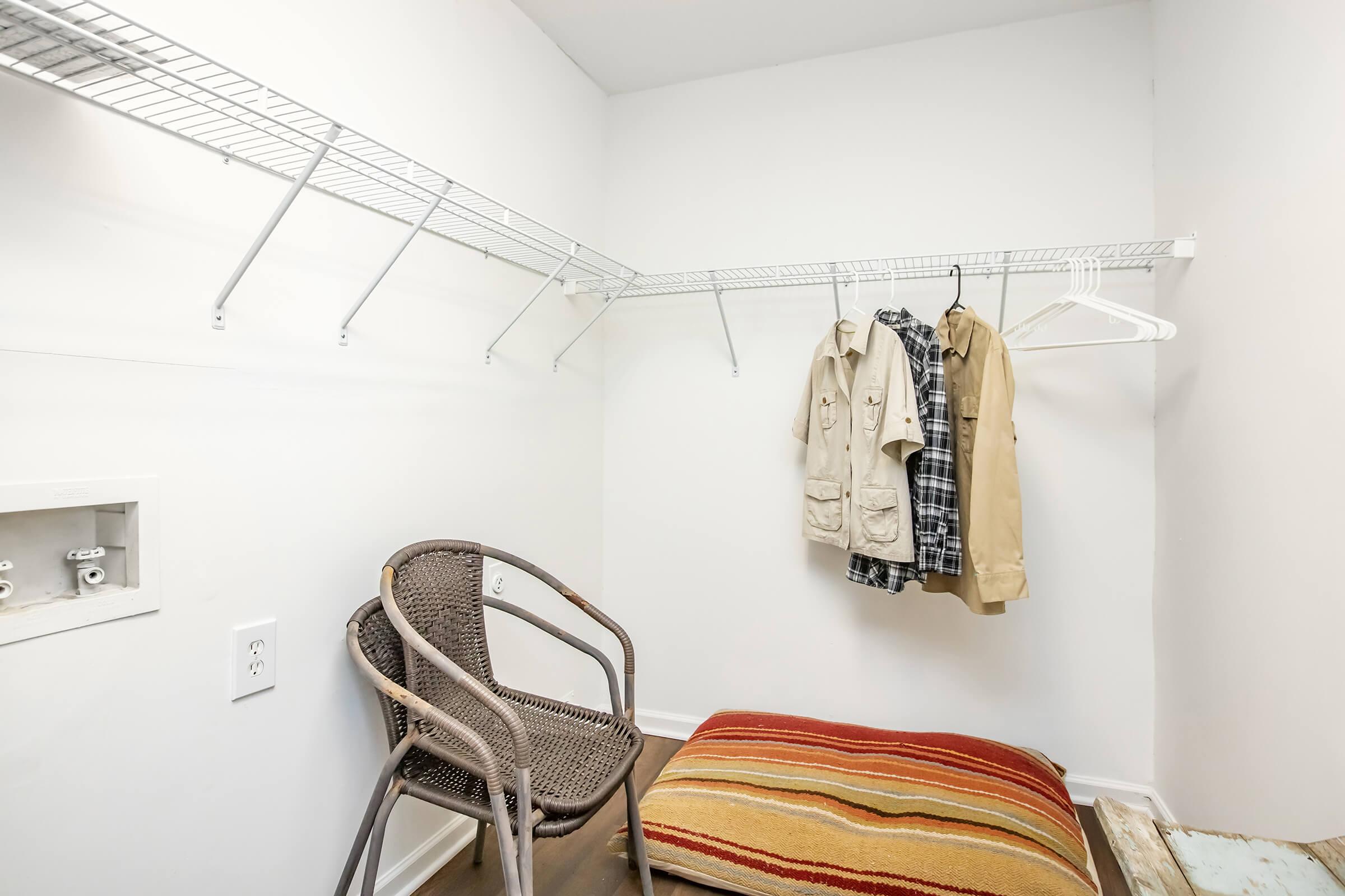 A small empty closet featuring a wire shelf with two hangers holding a beige jacket and a plaid shirt. There is a wicker chair in the corner and a striped mat on the floor, with white walls and a simple, clean appearance.
