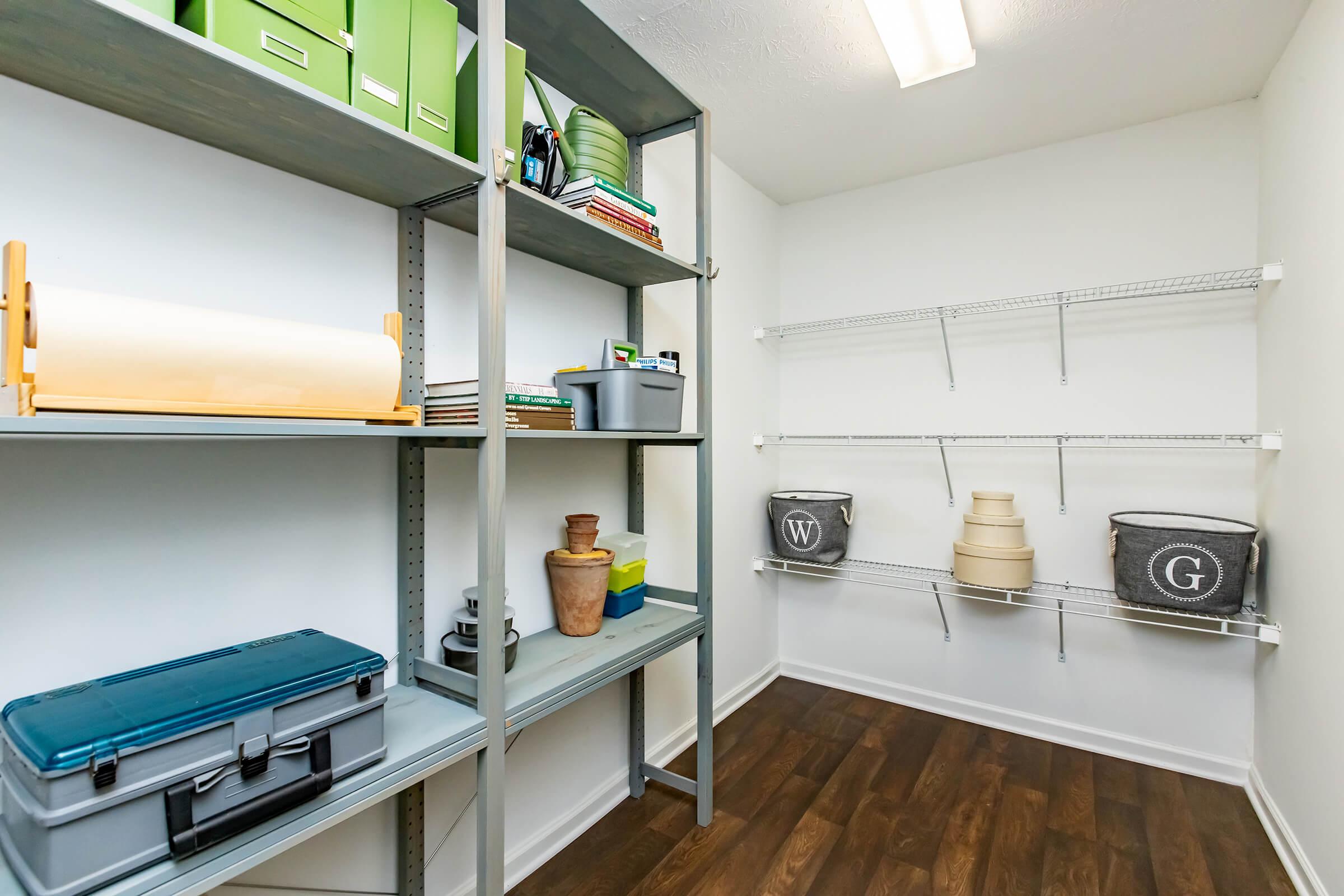 A well-organized storage area featuring shelving units with various items, including books, green storage boxes, a planter, and gray fabric bins labeled 'W' and 'G.' The walls are painted white, and the floor has a dark wood finish, enhancing the tidy and functional feel of the space.