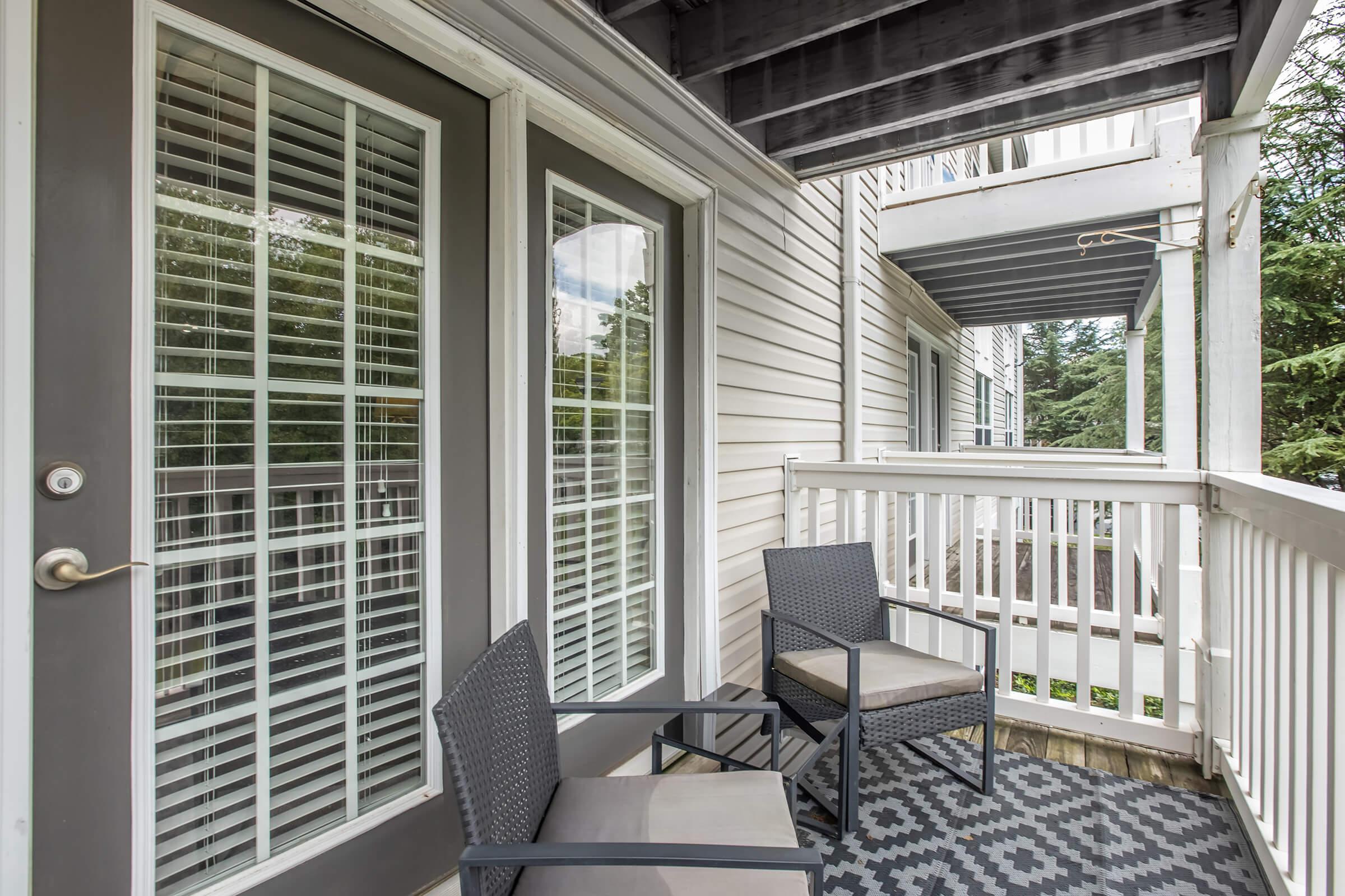 A small balcony featuring two black chairs with cushions, a patterned outdoor rug, and large glass doors with grid-pane windows. The balcony is surrounded by a white railing and has trees visible in the background, suggesting a serene outdoor space.