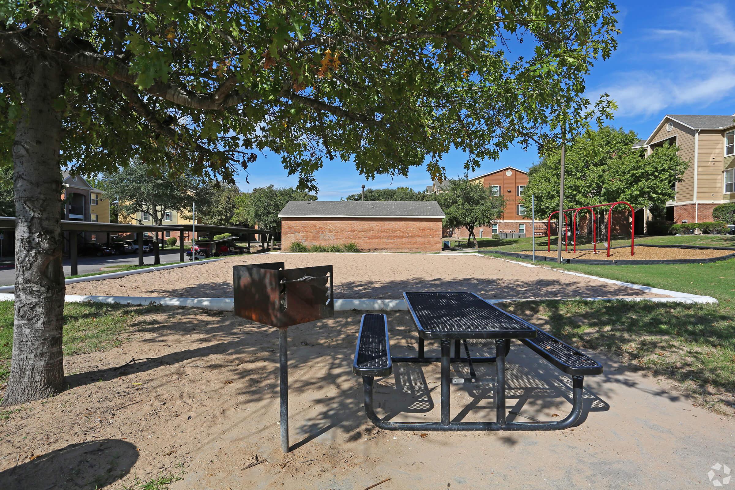 A picnic table with attached benches sits on sandy ground in a park. In the background, there is a volleyball court and playground equipment. The area is shaded by a large tree, with nearby apartment buildings visible against a clear blue sky.