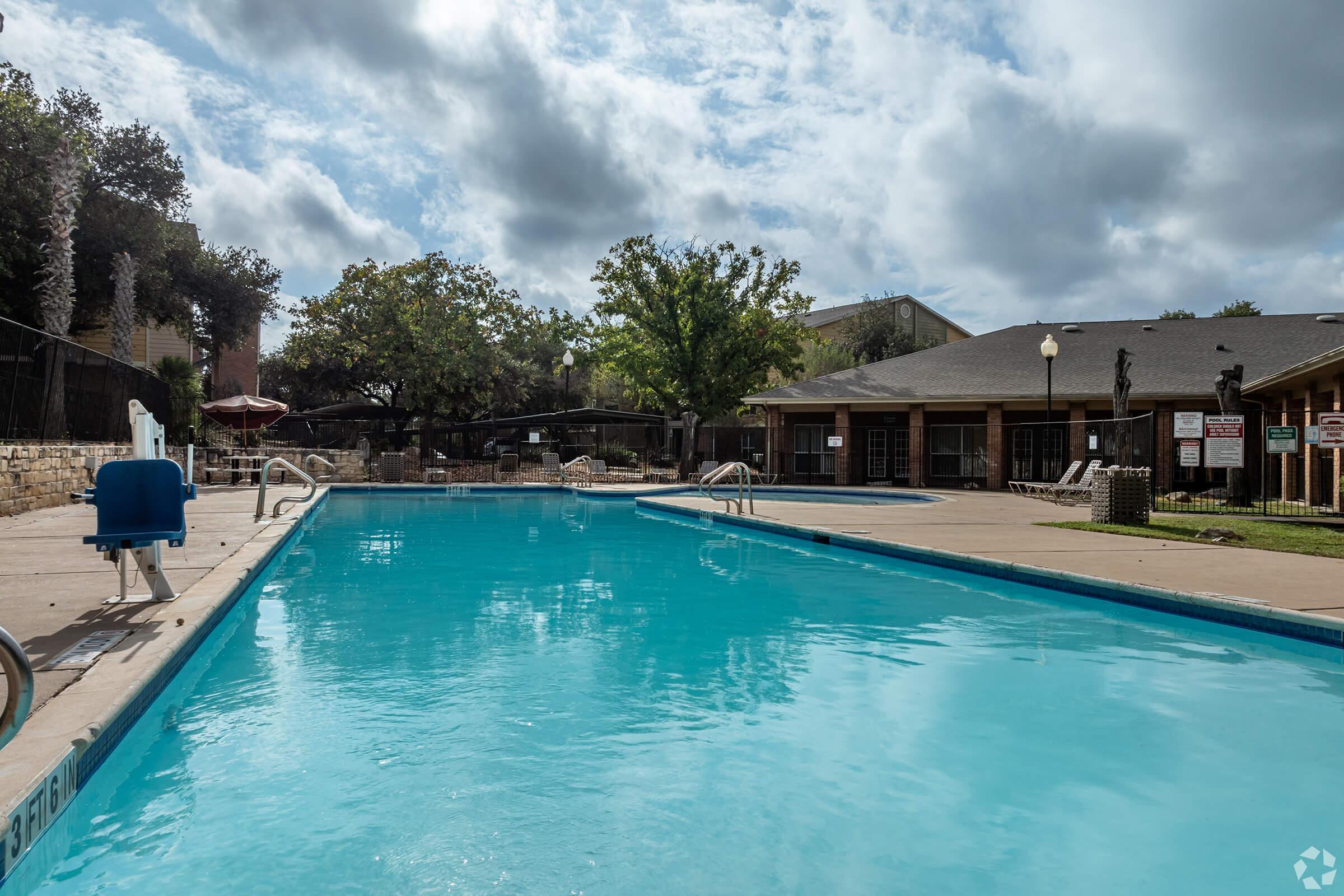 A clear, inviting swimming pool with bright blue water, surrounded by a concrete deck. In the background, a residential building with a patio area, trees, and a few poolside chairs. The sky is partly cloudy, creating a peaceful outdoor atmosphere.